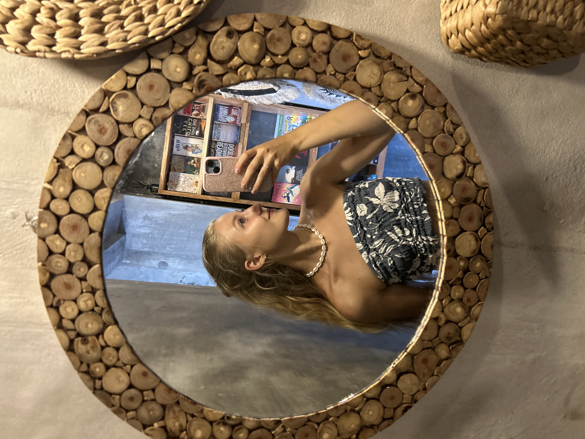 The image shows a reflection in a circular mirror with a frame made of small, round pieces of wood, possibly small tree trunk cross-sections. The reflection shows a woman holding up a cell phone, presumably to take a picture of herself in the mirror. She is wearing a strapless top with a floral pattern, and a necklace made of small, round shells.
Behind the woman in the reflection is a wooden shelf filled with books. A woven, straw-like item is seen in the upper right corner of the image and another similar item in the upper left. The wall behind the mirror appears to be painted in a light grey or off-white color.
The books and the woman's attire are consistent with a tourist environment, possibly in Sukawati, Indonesia as indicated in the source data. The environment has a natural, bohemian feel with the straw decor items.