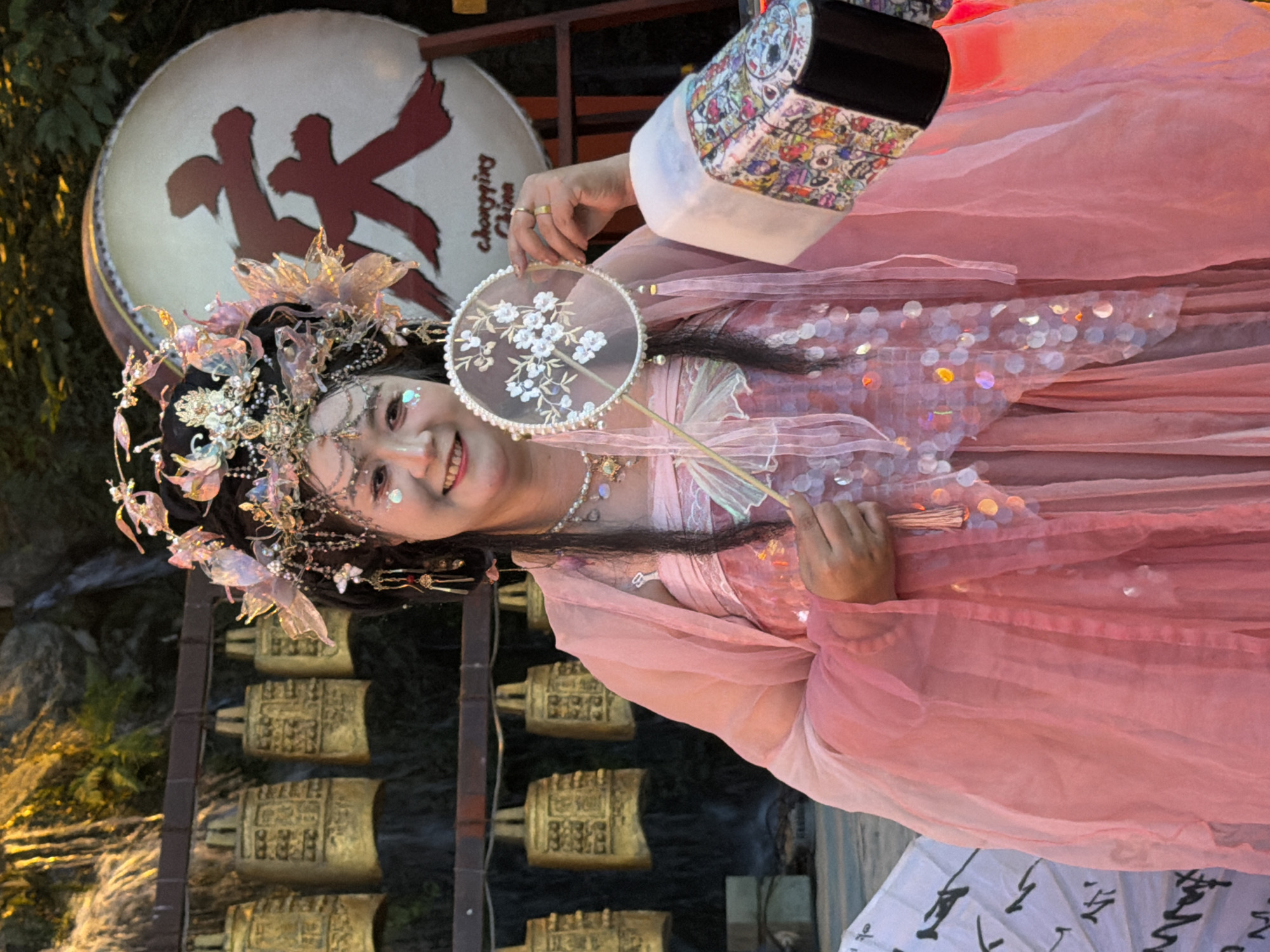 A young woman dressed in traditional Chinese attire, likely a Hanfu, poses for a photo. She wears an ornate headdress adorned with intricate floral and jeweled elements, and her pink dress is layered with sheer fabrics and embellished with sequins. She holds a small, circular fan decorated with a floral pattern. Her makeup includes glitter applied to her eyes and cheeks, and she wears delicate jewelry around her neck and fingers.
In the background, a large, circular drum with a prominent red Chinese character, "山" (Shān, meaning mountain) is visible. Below the drum, a series of golden bells hang in a row on a dark-colored stand, suggestive of a temple or cultural exhibit. The setting appears to be outdoors, with hints of greenery and possibly water features in the background, and the lighting suggests it might be late afternoon or early evening. The overall mood is festive and cultural, with the woman exuding a cheerful and engaging expression. The text "Chongqing China" is also subtly visible on the drum.
