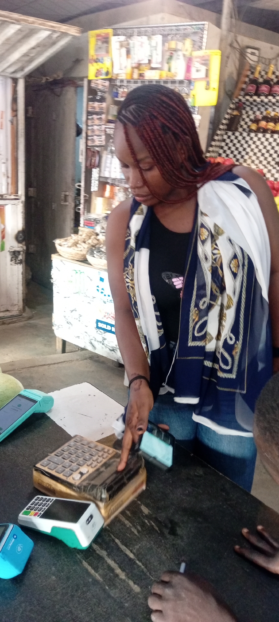 In Aviation Quarters, Nigeria, a young woman with braided hair, wearing a dark sleeveless top and a scarf with a white, blue, and gold patterned design, is engaged in a transaction at a market stall. She is leaning over a dark countertop, her right hand extended, touching a small, blue credit card reader. In front of her on the counter are two other payment devices: a larger, older calculator-style terminal with a gold casing and a smaller, modern white and teal credit card reader. Behind her, shelves are stocked with various goods, including bottles of what appear to be beverages and packaged items. The environment suggests an outdoor or semi-enclosed market setting with a focus on retail.