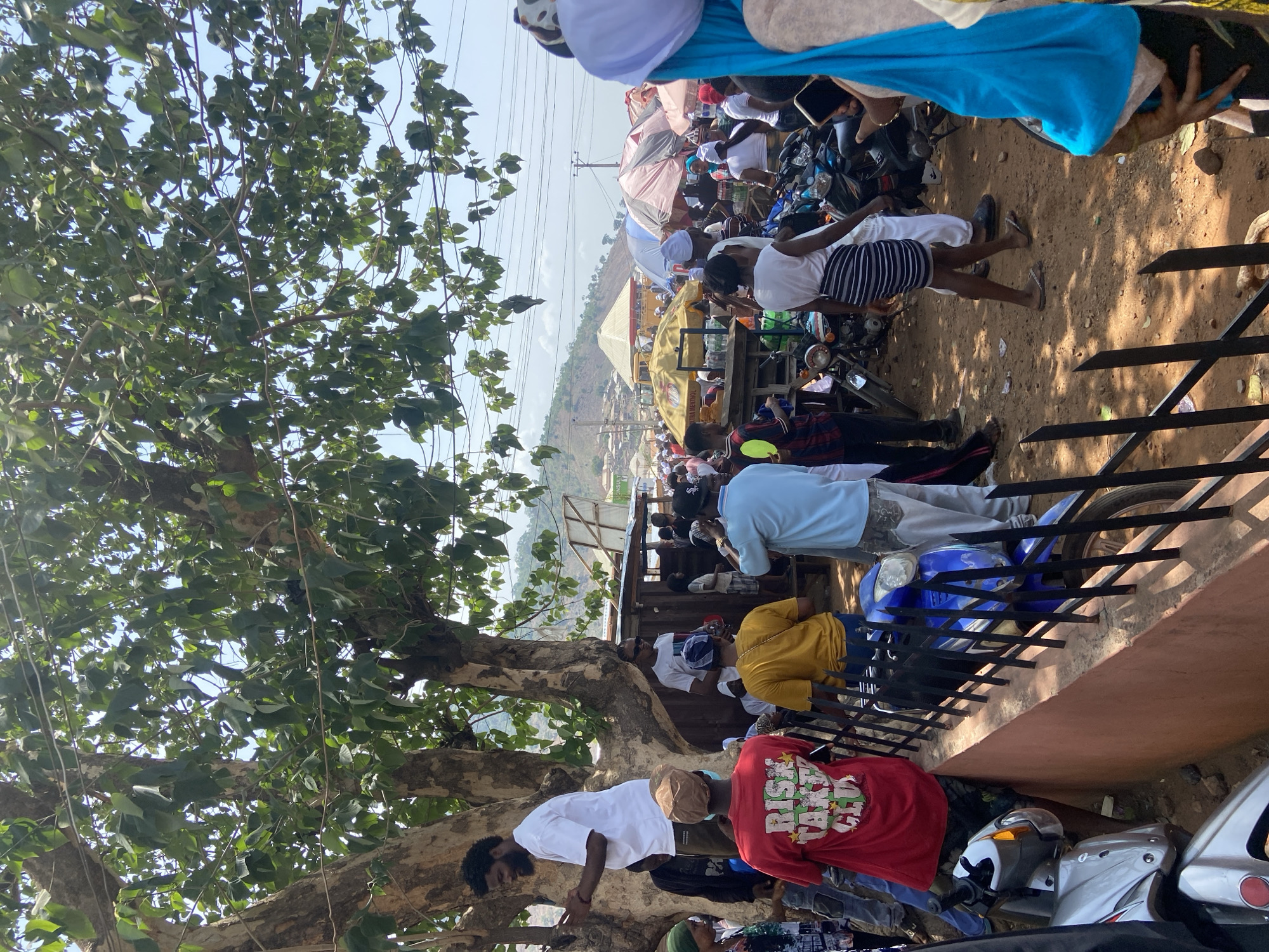 The image depicts an outdoor scene in Lokoja, Nigeria, possibly a market area. A large tree with dense foliage dominates the left side of the image. People are gathered in the middle and upper right areas, some wearing white clothing or head coverings, suggesting a religious event or festival. Motorcycles are parked among the crowd. A mountain is visible in the background. A fence runs along the right side of the image.