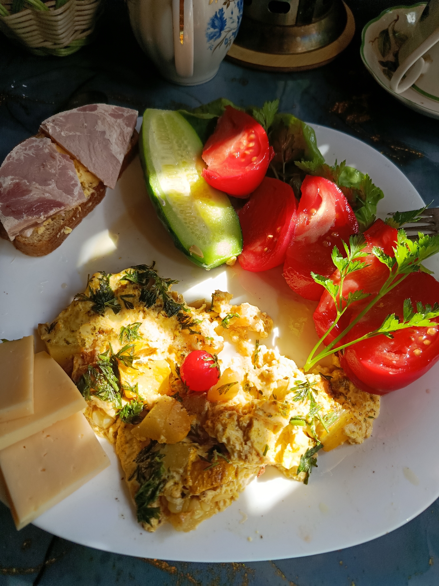 A close-up, overhead shot shows a breakfast plate filled with food. The plate contains a folded omelet topped with dill and a single cherry tomato, three slices of yellow cheese, a slice of bread with ham and butter, sliced cucumber, and sliced tomatoes with parsley. The plate rests on a dark blue, possibly marble, surface. In the background, a wicker basket and a white teacup with blue floral designs are visible, along with a brass coaster and another teacup. The lighting suggests natural sunlight is illuminating the scene.