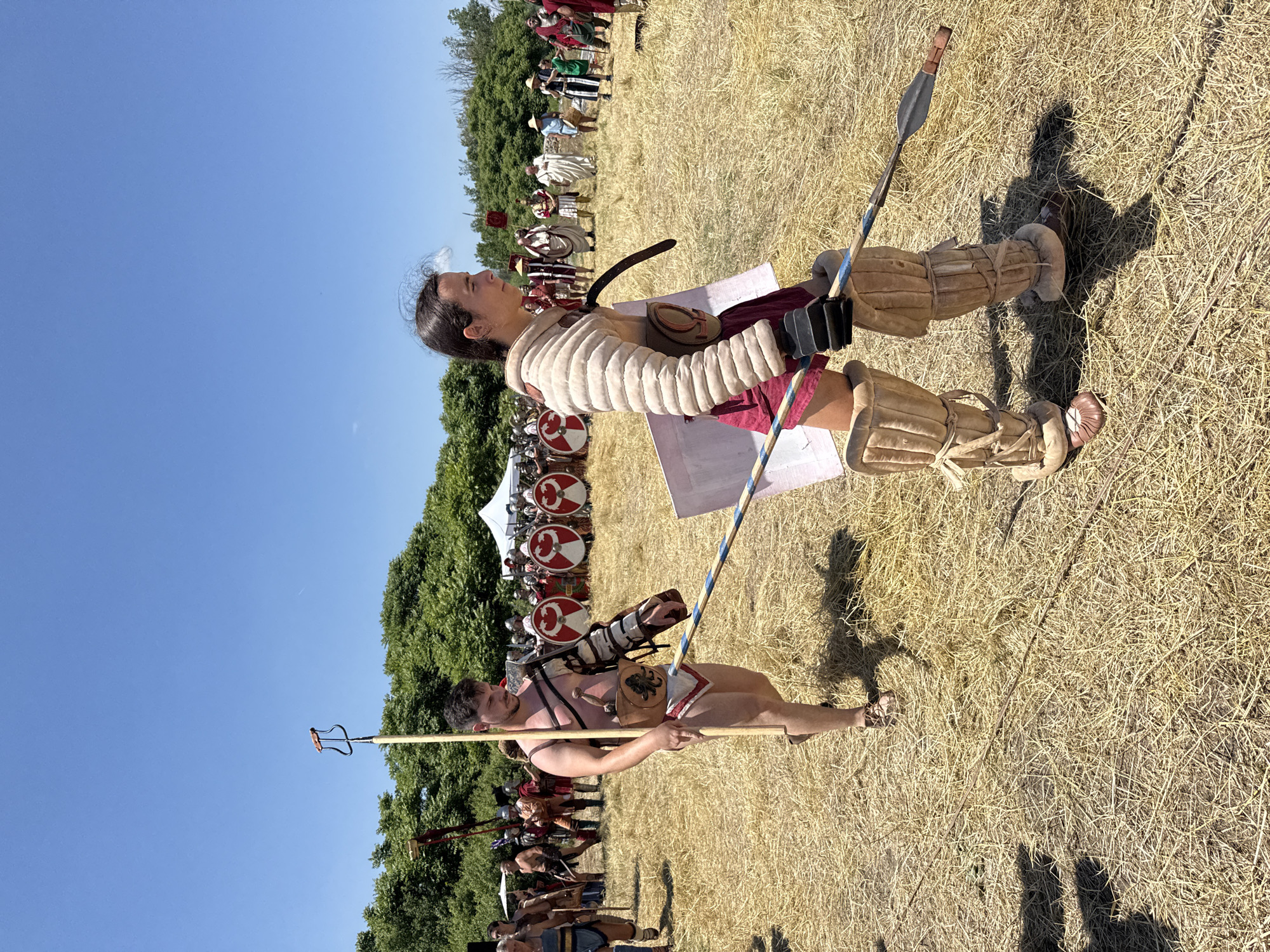This image captures a historical re-enactment scene under a clear blue sky in Gigen, Bulgaria, on a bright, sunny day. The ground is covered with dry, golden-brown straw and sparse dirt.
In the foreground, two individuals dressed as gladiators walk left-to-right across the frame.
The figure on the left is a male with dark hair and a short beard, bare-chested and wearing a white loincloth (subligaculum) with a red trim. He has a leather arm guard (manica) on his left arm, secured with straps, and a metal shoulder guard (galerus). A leather belt with a dagger in a pouch, embossed with a dark, octopus-like emblem, is strapped to his waist. He wears dark sandal-like footwear. He carries a long, light-colored wooden pole vertically in his right hand, topped with a distinctive dark metal, U-shaped implement with a wooden grip. His shadow is cast to his right.
The figure on the right is a younger individual with dark hair tied back, wearing a red garment (possibly shorts or a short tunic) underneath substantial padded light-tan leg guards (ocreae) and a padded arm guard (manica) on their right arm. They also wear dark sandal-like footwear. A white rectangular shield (scutum) is held against their left side, and a spear with a dark metal head is held diagonally, with the butt resting near their left foot and the shaft supported by the shield. This figure's head is turned slightly to the left, observing the male figure. Their shadow is cast to their right.
In the mid-ground, behind the foreground figures, a group of historical re-enactors is visible. To the left, several figures in various Roman-era clothing walk, one in a blue tunic, another in armor carrying a red standard. To the right, a line of Roman soldiers stands in formation, holding large round shields (scuta) decorated with concentric red and white circles.
The background features a dense line of lush green trees, above which a clear, cloudless blue sky extends. A faint white contrail from an aircraft is visible in the upper right quadrant of the sky. White tents are also present among the trees on the right side of the frame.