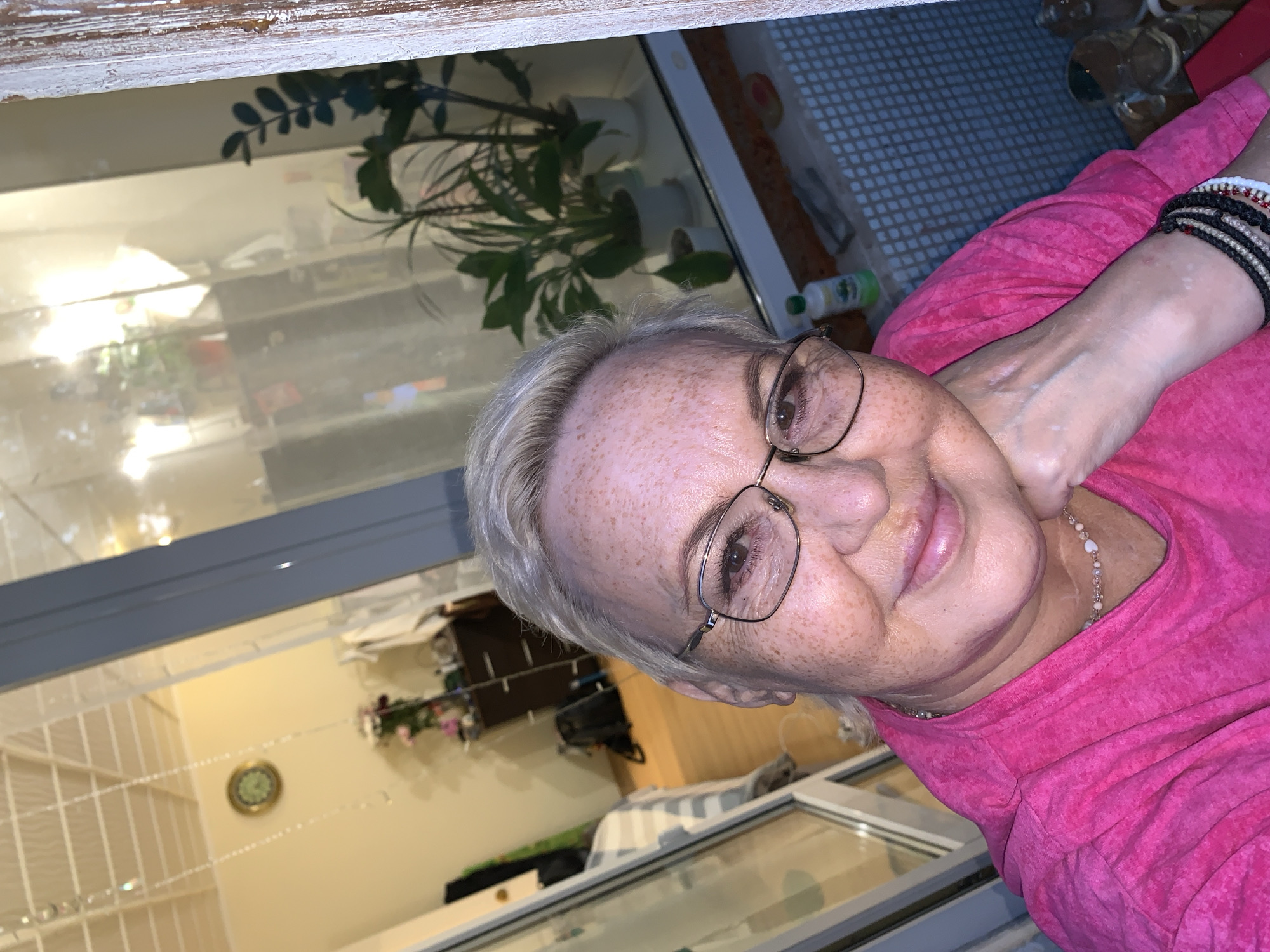 A close-up selfie shows a white woman with fair skin, freckles, and blonde hair cut short. She is wearing glasses with thin metal frames and a bright pink hooded sweatshirt. Her right hand is resting on her chin, and she is wearing several bracelets on her left wrist. In the background, through a window or glass door, a bedroom with a bed and a dresser with flowers can be seen. The overall impression is of a casual, indoor setting, possibly a home. There are no specific visual cues that would allow for an accurate determination of the location as Chaikovsky, Russia.