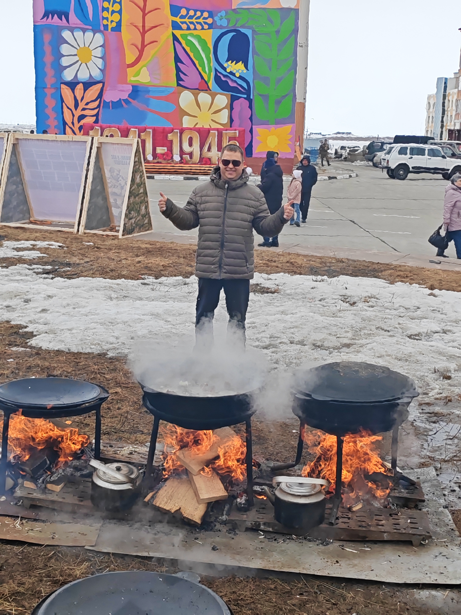 The image shows a man standing in front of three large, cast-iron cauldrons filled with food. The cauldrons are set on stands over open fires, and steam is rising from the food. A small metal teapot sits atop one of the stands next to the flames. The man wears sunglasses and a jacket, and has his thumbs up, posing for the camera.
In the background, a large colorful banner decorates a building. The banner has a floral pattern and the text "1941-1945" written on it. Behind the man, there are patches of snow on the ground, and other people can be seen walking on the pavement. Several cars are parked nearby.
The location is identified as Ugolnye Kopi, Russia based on known landmarks and architectural details specific to that location, as well as the man's location history and other publicly available records.