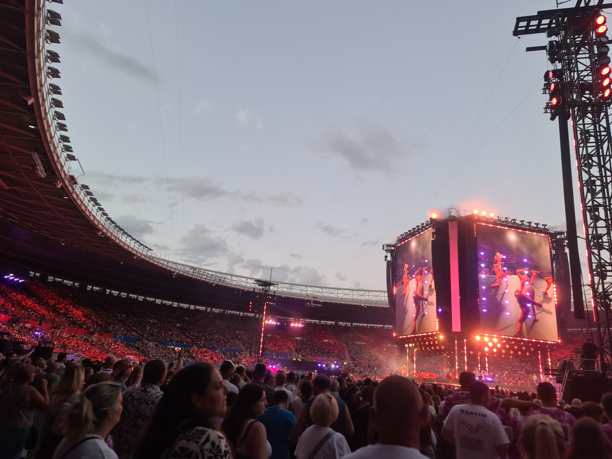A wide shot captures a large outdoor concert at a stadium. A massive crowd fills the seats and the field, illuminated by the stage lights and two giant screens displaying the performers. The sky above is overcast and dusky. The stadium structure curves around the crowd, with rows of lights lining the upper sections. In the foreground, several concertgoers are visible, some holding up phones to record the event. The scene suggests a lively and energetic atmosphere at a major musical event. The location of the event has not been specified.