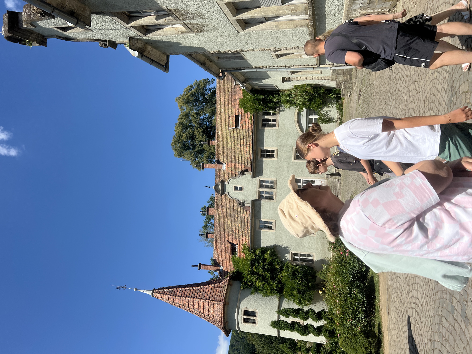 The image shows a group of people walking in the courtyard of a castle in Karpaty, Ukraine. The castle has a tall, conical tower with a red tiled roof and a weather vane. The walls of the castle are a pale green color, and there are several windows and chimneys visible. The courtyard is paved with cobblestones. In the foreground, a woman wearing a white and pink checkered t-shirt and a wide-brimmed hat is walking towards the left. To her right, a teenage girl in a white t-shirt and green shorts is walking away from the camera. Behind them, a young boy in a black t-shirt and black shorts is walking. To the far right, a man in a black t-shirt and black shorts is walking away from the camera. The sky is a clear blue with a few white clouds. The environment is sunny.
