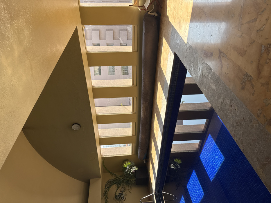 An indoor swimming pool facility in an urban area of Mexico City, Mexico, is depicted under natural light. The scene focuses on a rectangular dark blue tiled swimming pool, featuring a silver metallic ladder on its left side. Sunlight streams through a series of five vertical, rectangular windows on the far wall, casting distinct, elongated rectangular shadows on the light brown tiled floor bordering the pool. The water's surface reflects these vertical window frames and the bright sky.
Along the base of the windows, a large, dark beige or gray rolled-up pool cover is horizontally positioned. To the far right, a white folding chair is partially visible. A small, round wall clock with a white face and a dark frame is mounted on the wall above the windows. In the far left corner, a large potted palm-like plant with dark green leaves stands in a square, dark-colored planter. The walls are a uniform light beige, leading up to a curvilinear, arched ceiling painted in a lighter yellowish-beige. Through the windows, views of other multi-story urban buildings with light-colored façades are visible. No individuals are present in the image.