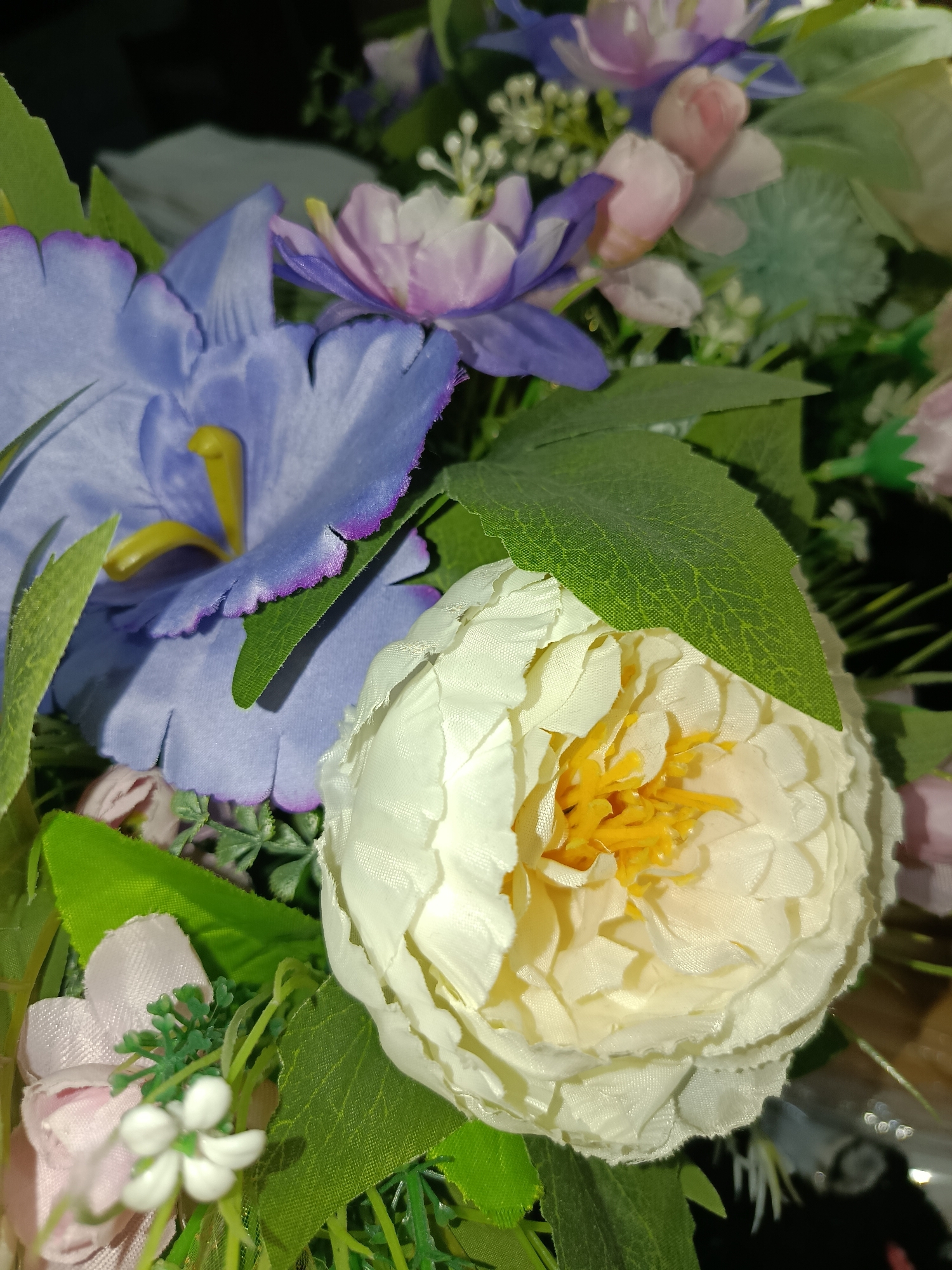 The image displays a close-up view of a bouquet of artificial flowers, predominantly featuring a large white peony in the foreground, with its yellow stamen visible. To the left, a large purple flower with a yellow center and a ruffled edge is partially visible. Other flowers and greenery, including pink and light purple blossoms and green leaves, are interspersed throughout the arrangement. The lighting suggests an indoor setting, possibly a room or a decorated space. The scene does not provide any specific geographical or locational context that would allow for identification within Tashkent, Uzbekistan, or any other city. There are no people or actions depicted in the image.
