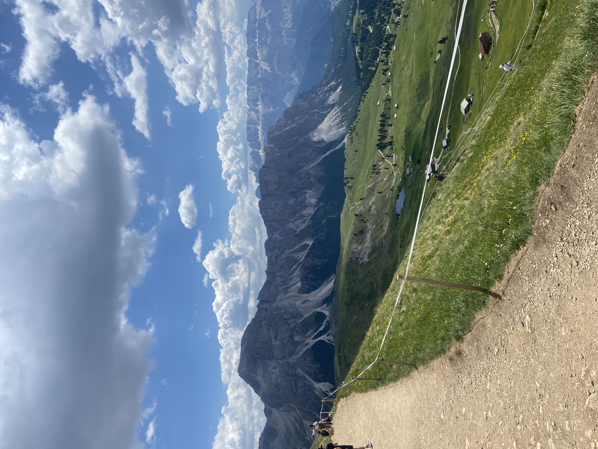 The image depicts a mountainous landscape under a partly cloudy sky. The foreground features a dirt path leading along a grassy slope. A white rope barrier, supported by wooden posts, runs along the edge of the path. Several people are visible on the path and near the barrier, some appearing to be hikers.
In the mid-ground, a vast green meadow stretches out, dotted with small buildings, possibly chalets or mountain huts, and scattered trees. A small, dark body of water, likely a lake, is visible in the distance. Beyond the meadow, a dramatic, rugged mountain range dominates the scene. The mountains are characterized by steep, rocky slopes, with patches of snow visible on some of the higher peaks. The scale of the mountains suggests they are part of a significant mountain range.
The sky is a mixture of bright blue and white clouds, some of which are large and fluffy, while others are more wispy. The sunlight suggests it is daytime, likely late morning or afternoon. The overall impression is of a scenic and expansive outdoor environment, possibly a popular hiking or tourist destination. The clear weather and the presence of people enjoying the outdoors evoke a sense of leisure and exploration. There is no visible text in the image.