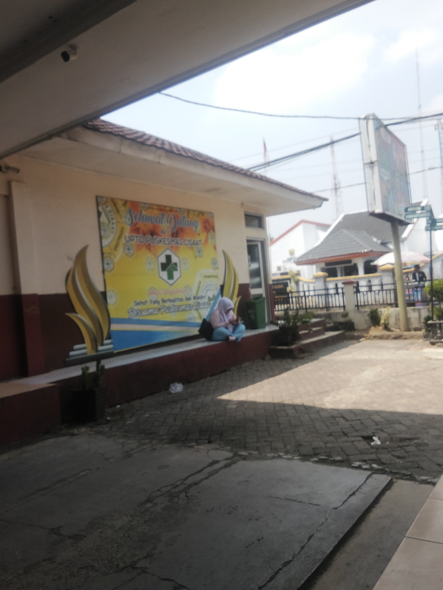 The image depicts the exterior of the UPTD PUSKESMAS CISAA'T, a public health center in Cisaat, Indonesia. A banner with "Selamat Datang" (Welcome) is prominently displayed on the building's facade, featuring a green cross emblem. Two individuals are present: one is sitting on the ground near the banner, and another person is standing in the background near a white fence and umbrellas. The foreground shows a paved area with some debris, including a crumpled white plastic bag. The background reveals several buildings, including a white structure with a grey roof, and telecommunication towers in the distance. The sky is overcast.