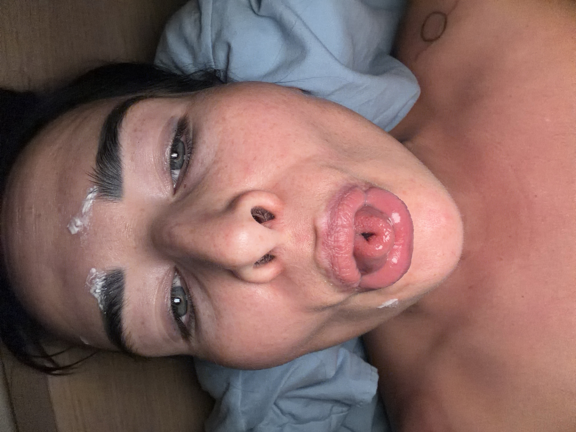 The image shows a person lying down with their face looking upwards. The person has black hair and grey eyes. There is a white substance on their left eyebrow and the corner of their mouth. Their tongue is sticking out. There is a blue cloth under their head and shoulder. The image provides no information about the location being in Saint Petersburg, Russia.