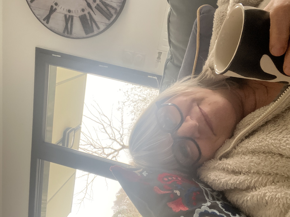 This image depicts a middle-aged woman reclining indoors in Chambœuf, France. The woman has shoulder-length, light-colored hair and wears dark-rimmed glasses, presenting a subtle smile. She is wearing a light-colored, textured, possibly fleece, zip-up garment. Her head rests on a dark-colored pillow adorned with red, blue, and white embroidered patterns. In her right hand, she holds a black and white mug with an organic design. The woman appears to be seated or lying on a dark-colored sofa. In the background, a large window reveals an outdoor scene with bare trees and a light-colored outdoor umbrella. To the upper right, a large, round wall clock with Roman numerals and a distressed white face is visible on a light wall. The lighting suggests ambient daylight, with the outdoor view appearing bright.