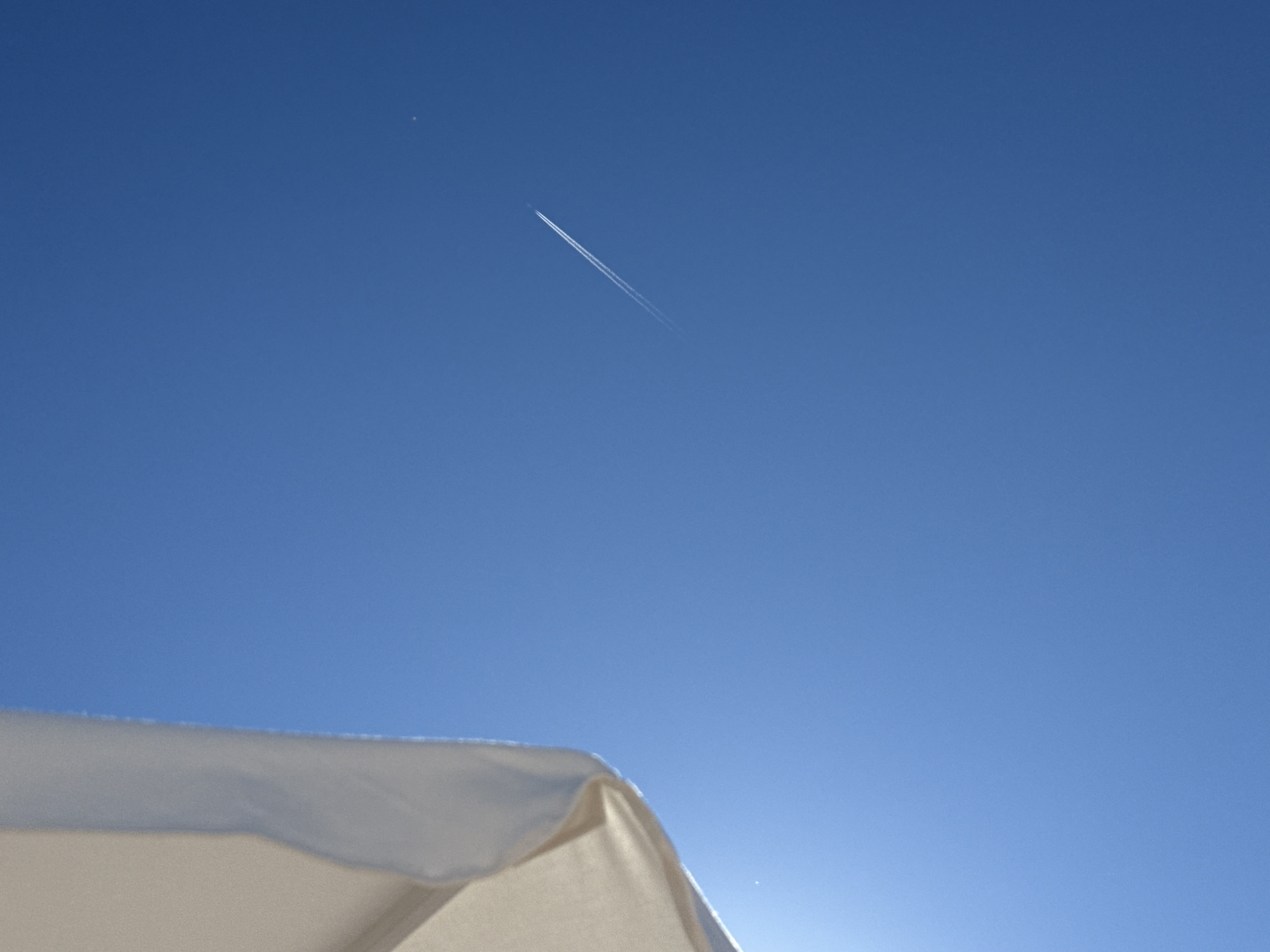A low-angle, wide shot captures a bright blue sky, possibly over Sozopol, Bulgaria. In the upper left quadrant, a portion of a light-colored, possibly beige or cream-colored, fabric object, such as a beach umbrella or awning, occupies the frame. This object is partially illuminated by sunlight, creating a bright, washed-out area at its edge. Across the vast expanse of the sky, a thin, white contrail from an airplane stretches diagonally from the middle left towards the bottom right. A small, faint, white dot, possibly a distant star or the moon, is visible in the upper right quadrant of the sky. The scene suggests a clear, sunny day.