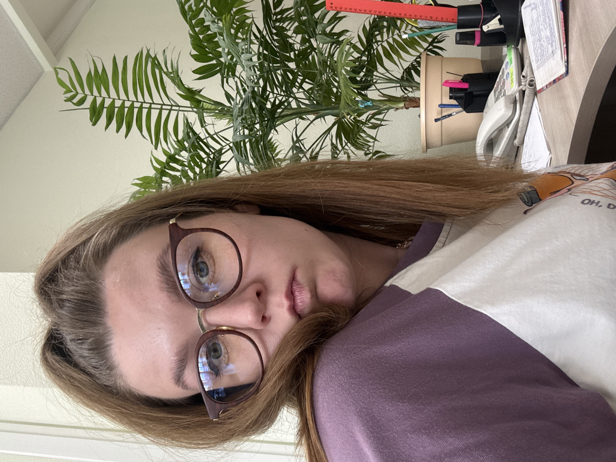 An indoor, rotated portrait shows a young woman looking directly at the camera. Her head is positioned on the left side of the frame, and her body extends towards the right. She has light brown hair, which is slightly pulled back, and wears reddish-brown framed eyeglasses that reflect ambient light. Her eyes are blue or green, and her expression is neutral and direct, with her lips slightly parted. She is wearing a two-toned top, primarily dark purple/mauve and white, with the partial text "OH, D" visible on the white section of her shirt.
Behind her, a light-colored wall serves as the background. To the upper left, a portion of a white ceiling with a recessed panel is visible. A large, artificial-looking green plant with numerous palm-like fronds in a light brown pot occupies much of the background space, suggesting an indoor setting like an office or study.
On a light-colored desk to the right, several objects are arranged: a white landline phone with a coiled cord, a black stapler, and various pens or pencils in a black holder. A red ruler with visible numerical markings (e.g., "20" through "30") rests near the plant. A notebook or binder with illegible handwritten text is also on the desk. The image appears to be a close-up selfie, offering a glimpse into an everyday indoor environment, potentially in Izhevsk, Russia.