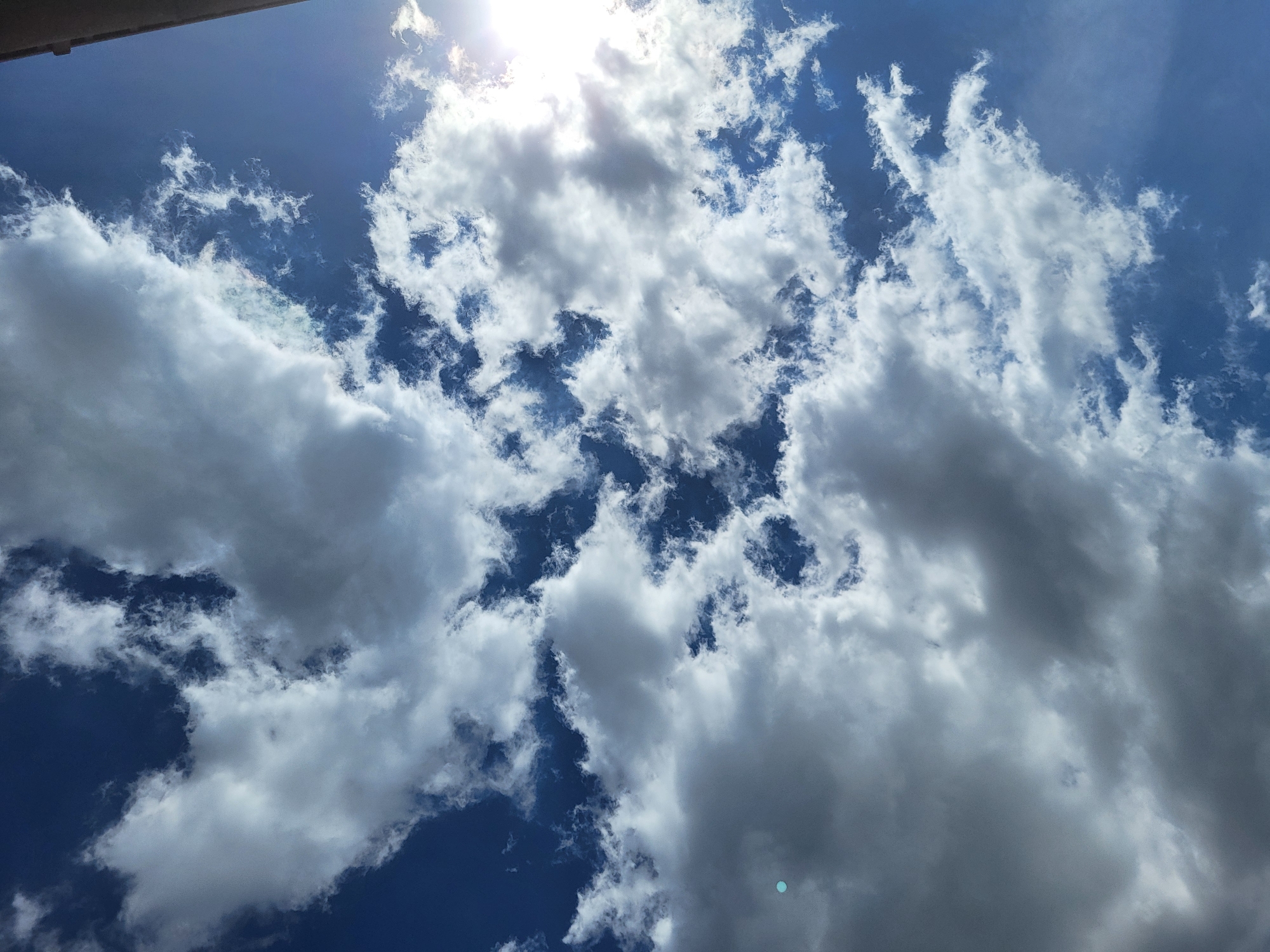 The image is a low-angle shot of a bright, blue sky with numerous white clouds. The sun is shining brightly, partially obscured by some of the clouds at the top. The clouds vary in size and density, from small wispy formations to large, puffy ones. There is a small piece of what appears to be a building or structure visible in the very top left corner of the frame. The overall impression is that of a sunny day with a dynamic cloudscape. The location appears to be outdoor due to the presence of the sky and clouds. The city is not apparent from the image.