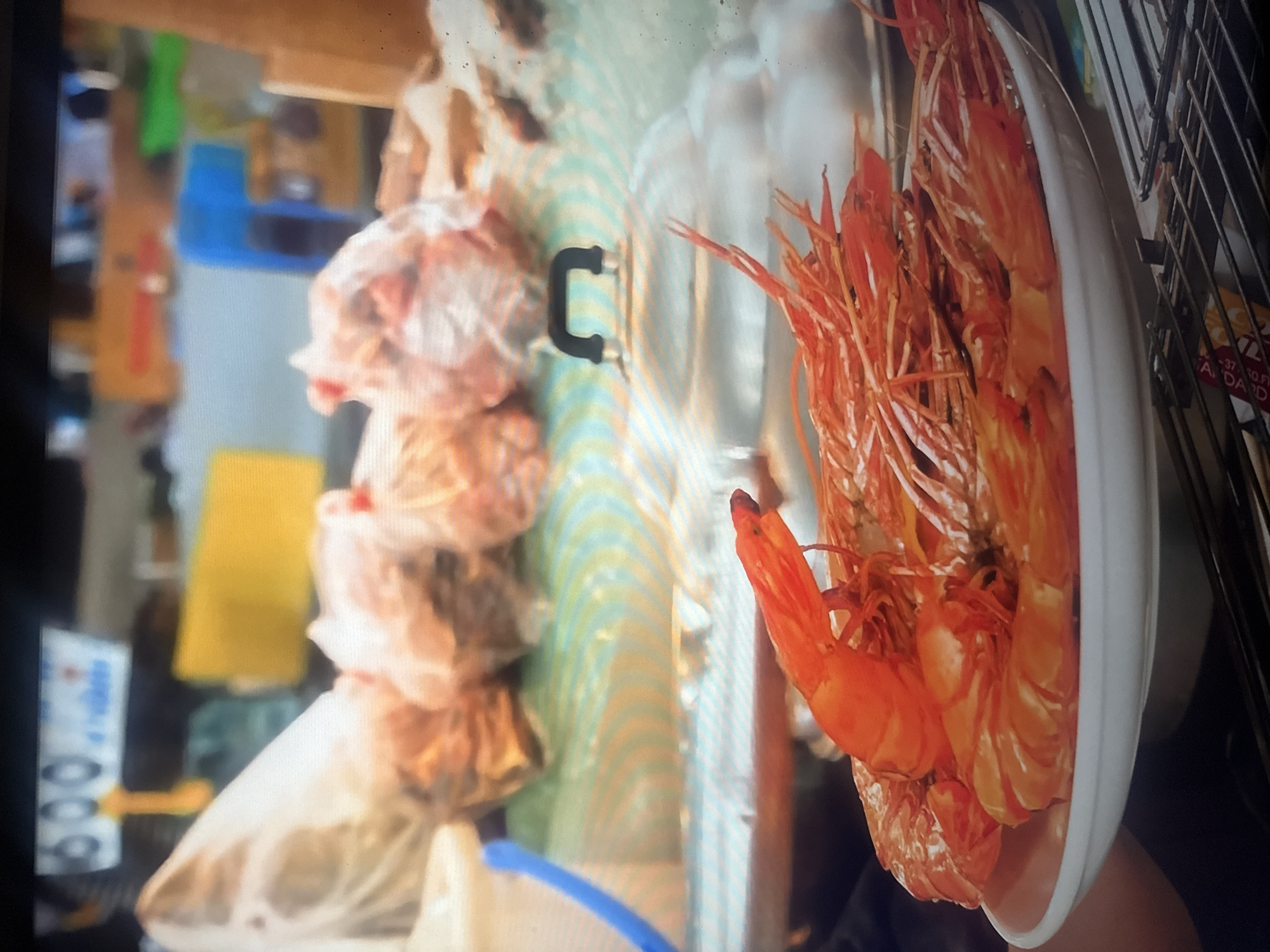 The image depicts a close-up view of several cooked shrimp in a white bowl, positioned in the foreground. The shrimp are bright orange and appear to be whole, with their heads and legs still attached. They are arranged in a way that fills most of the bowl, suggesting a generous portion.
In the background, a variety of objects are visible, though they are out of focus and less distinct. There are what appear to be plastic bags containing food items, possibly raw ingredients or prepared dishes, arranged vertically. Some of these bags have a slightly translucent quality, revealing hints of their contents. Further back, there are hints of a market stall or food preparation area, with shelves or racks and various containers. A yellow rectangular object and some blue and green elements are also present in the mid-ground, adding to the sense of a busy environment.
To the right of the bowl of shrimp, a metal grate or rack is visible, along with a small yellow and white sign. The sign has some text on it, but it is largely illegible due to the image quality and angle. However, a few letters and numbers, possibly a date or price, can be vaguely discerned. The overall impression is that of a food market or a culinary setting, possibly an outdoor stall or a kitchen counter, in Herzliya, Israel. The lighting suggests it could be daytime. No people are clearly visible in the frame.