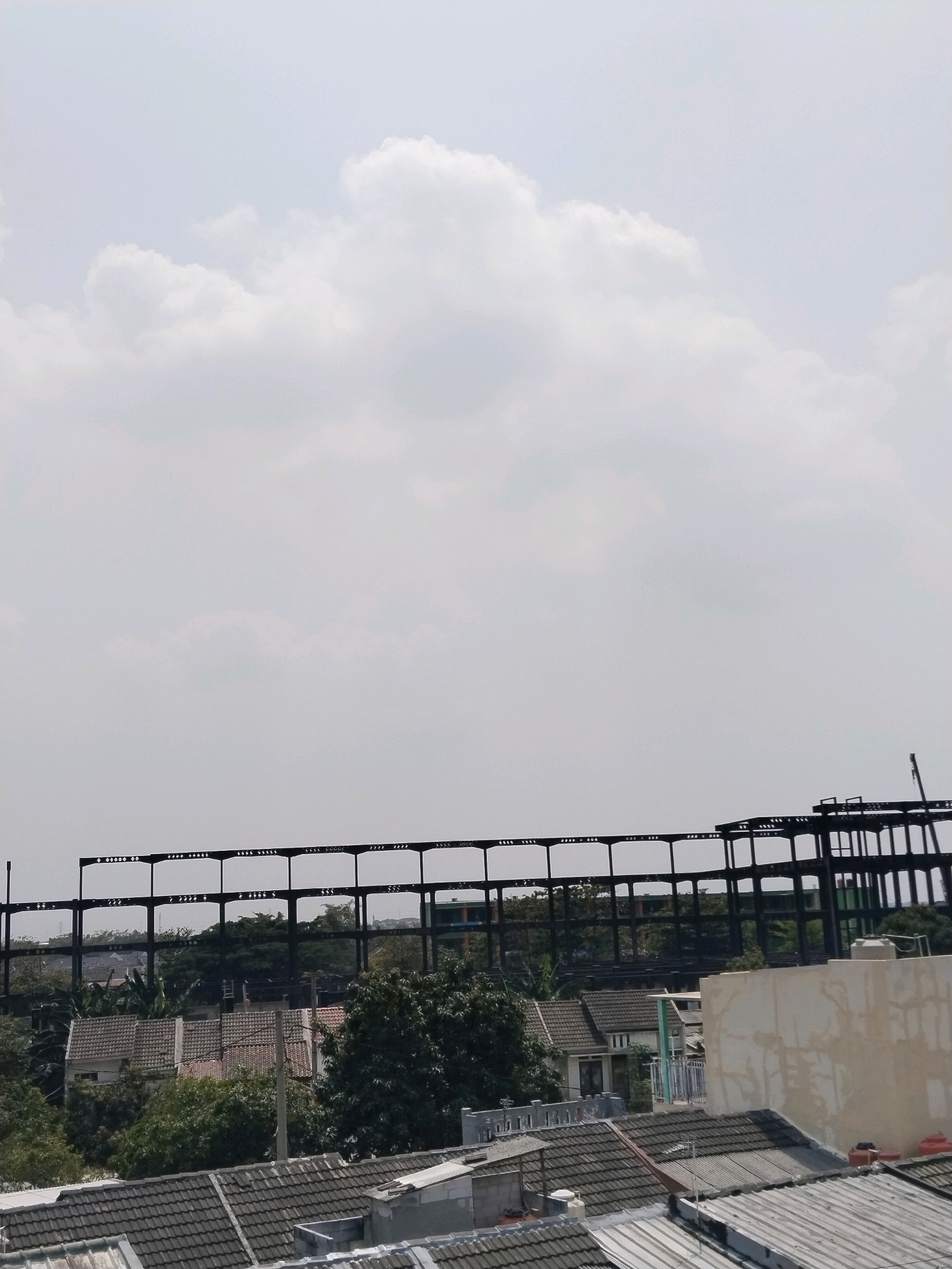 The image depicts a partially constructed building with a skeletal steel frame against a pale, overcast sky. Below the construction, a residential area is visible, characterized by tiled rooftops and lush green trees. The scene appears to be an urban or suburban environment in Jalenjaya, Indonesia, judging by the architectural style and vegetation. The overall impression is one of ongoing development within an established community. There are no people or animals present in the frame.