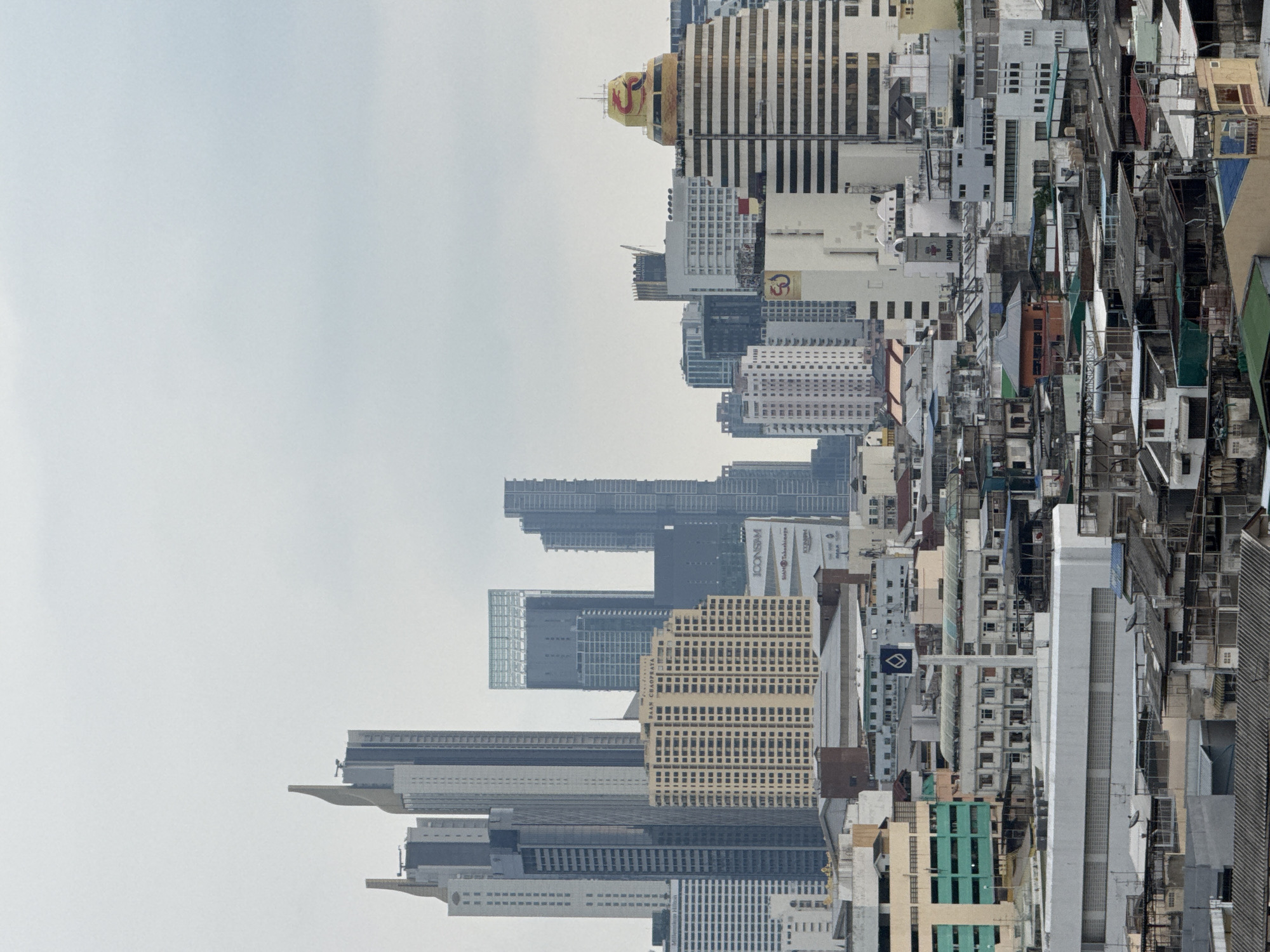 The image shows a cityscape of Bangkok, Thailand, viewed from an elevated position. The buildings are a mix of modern high-rise structures and older, low-rise constructions. The skyline is uneven, with varying heights and architectural styles. The sky is overcast, providing a muted backdrop to the urban scene. The image is rotated 180 degrees.