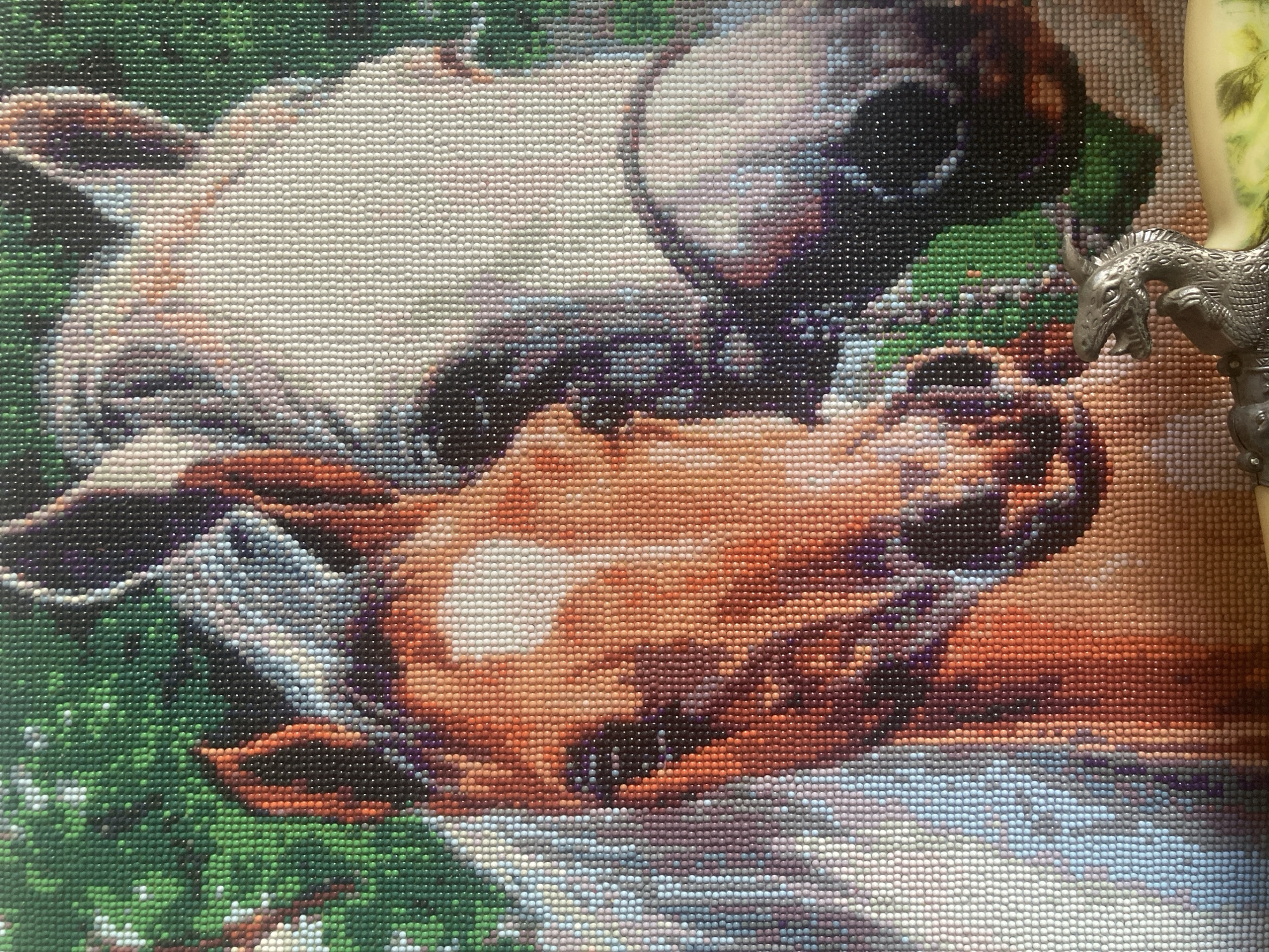 The image is a close-up, detailed view of a diamond painting depicting two horses. The horse in the foreground, on the left, is a lighter brown with a white blaze on its forehead, and appears to be facing slightly to the left. Its mane is white and gray. Behind it and to the right is another horse, primarily white, with its head turned more towards the viewer. The background is a mosaic of green dots, suggesting foliage. A small, metallic figurine of a dragon is visible in the bottom right corner of the frame, resting on an unseen surface. The image is a close-up of a handicraft, not a real-life scene. Therefore, it cannot be associated with a specific location such as the city of Marx, Russia.