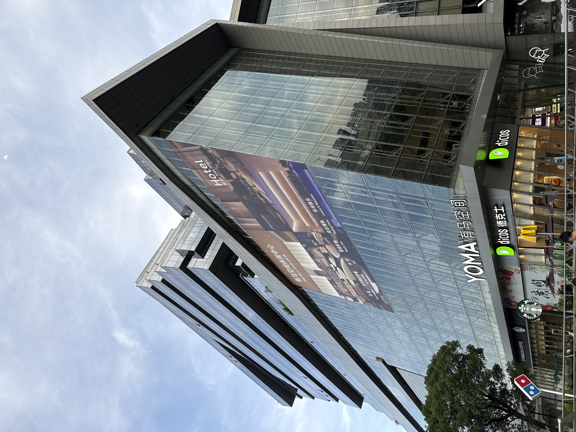 The image shows a tall, modern building with a glass facade, likely in Haizhu District, China, based on the Chinese writing and signage. A large banner advertising a hotel ("Hotel, 2F of this mall") is displayed on the glass. Several businesses are located at the base of the building, including YOMA Space, Dicos (a fast-food chain), Starbucks Coffee, and Domino's. The sky is visible above the building, with some clouds present. A tree is partially visible near the base of the building. Some light poles are visible in the foreground on the right side.