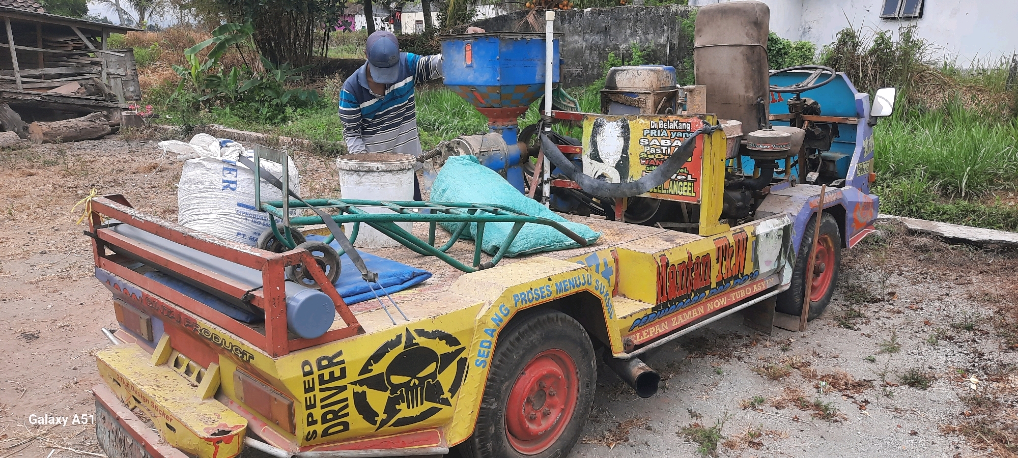 In Ploso, Indonesia, a yellow and blue modified vehicle, likely a small truck or tractor, is parked on a dirt and gravel surface. The vehicle is equipped with what appears to be a grain milling or processing apparatus, including a blue hopper, a brown tank, and an engine. A man wearing a blue cap and a striped long-sleeved shirt is operating the machine, interacting with a white bucket positioned beneath the hopper.
The vehicle's platform is laden with various items, including a large white bag with blue lettering, a turquoise-colored sack, and green metal bars. The sides of the vehicle are decorated with colorful graphics and text, including a skull and crossbones emblem with the words "SPEED DRIVER," and other Indonesian phrases.
The background consists of vegetation, trees, and a partially visible wooden structure on the left, suggesting a rural or semi-rural setting. The overall scene depicts an active agricultural or processing operation.