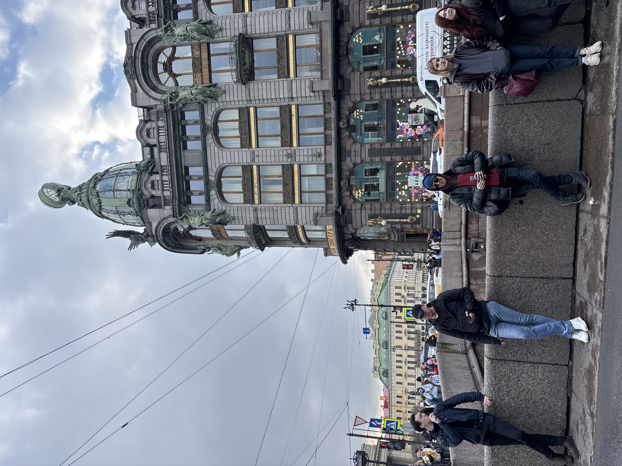 The image depicts Nevsky Prospect in Saint Petersburg, Russia, with a view of the Singer House. The Singer House is a prominent architectural landmark with a distinctive glass dome topped with a statue. Several people are sitting and lying along a stone embankment, apparently relaxing and taking in the scenery. They are dressed in casual clothing. A white van is parked near the Singer House. In the background, there are buildings and a crowd of people on the sidewalk. The sky is overcast. Power lines stretch across the sky. Traffic signs and street lamps are visible on the street.