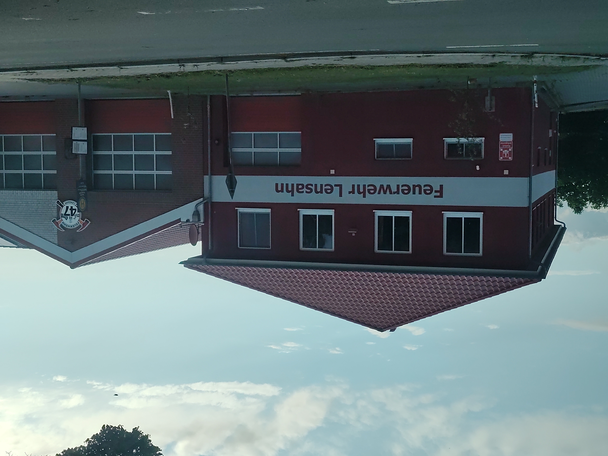 The image shows the fire station in Lensahn, Germany. The building is predominantly red with a red tiled roof. A large white band runs across the front of the building, bearing the text "FEUERWEHR LensaHN" in dark red lettering. The fire station has multiple windows and at least two large bay doors, one of which is visible on the right side of the building and appears to be a garage door with a grid pattern. To the right of the main building, there is an extension with a brick facade. Above the bay door, there is a circular emblem with the number "47" and the word "FEUERWEHR" (fire department) written on it. The building is situated next to a road, with a sidewalk and grassy area in front of it. Trees are visible in the background to the left of the building. The sky above is partly cloudy. There are no people visible in the image.
