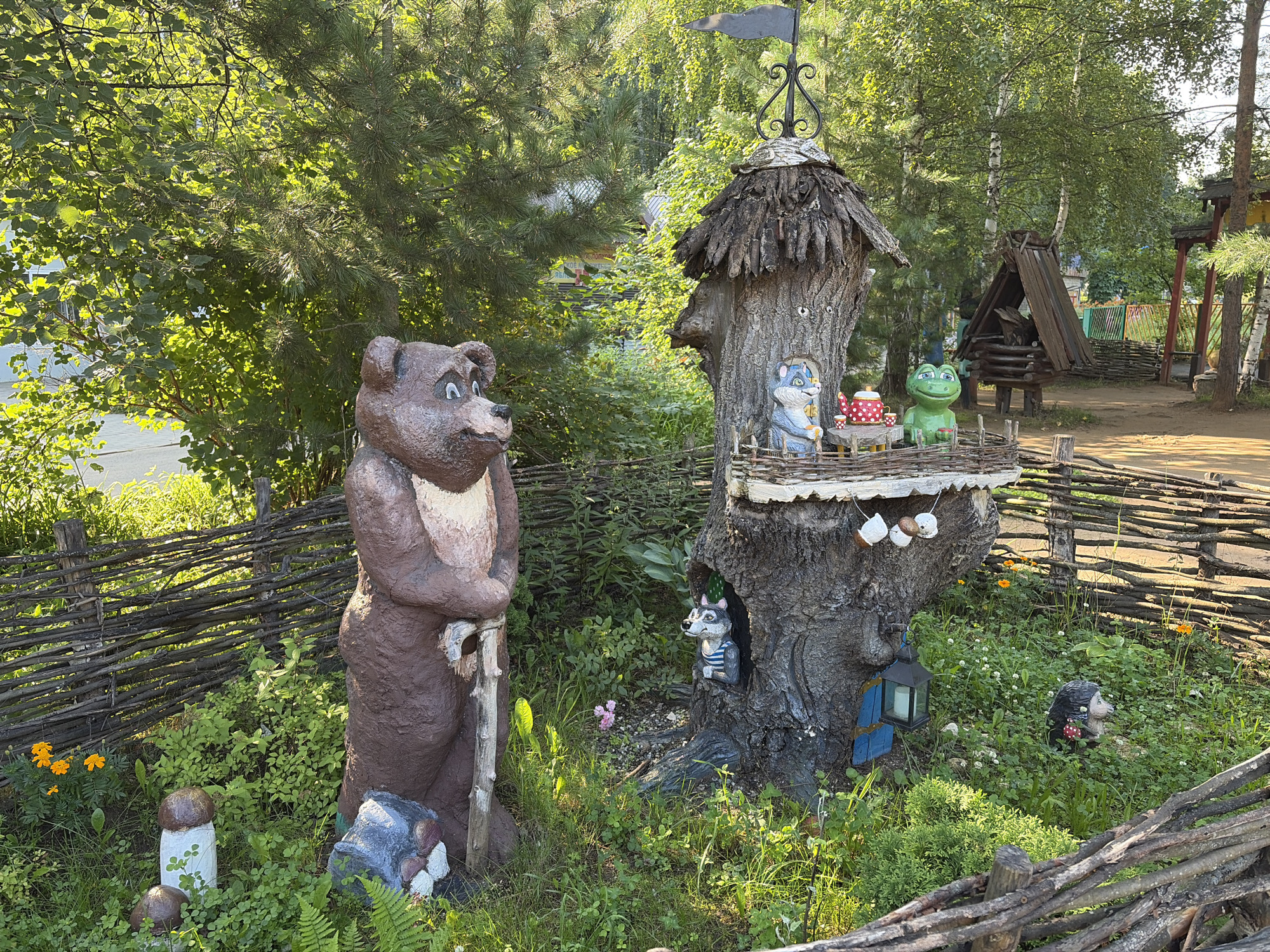 In the city of Chaikovsky, Russia, on a sunny day, an outdoor decorative display features several animal figurines amidst lush greenery and a rustic wooden fence. A large, brown bear statue stands prominently to the left, leaning on a wooden pole. To its right is a tree-like structure with multiple levels, adorned with a thatched roof and a weather vane. On the upper level, a blue mouse figure is seated at a small table with a red and white polka-dotted teapot and two cups. A green frog statue sits beside them. Below, a gray wolf figure wearing a striped shirt peeks out from an opening in the tree trunk. At the base of the tree structure, a black lantern with a candle inside is attached, and a small hedgehog figure with a red mushroom is nestled in the clover. In the foreground, two mushroom-shaped garden decorations and a grey rock with white and red painted details are visible. The environment is characterized by dense green foliage, scattered small orange and white flowers, and a rough-hewn wooden fence. In the background, wooden structures and more trees suggest a park or recreational area.
