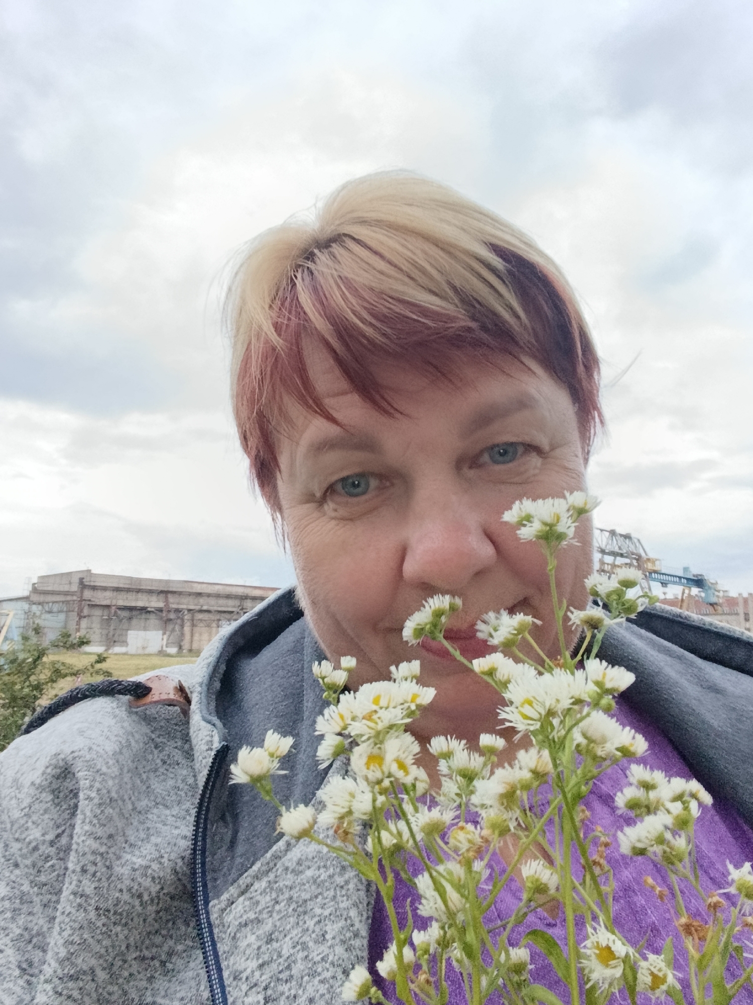 A selfie shows a middle-aged woman with blonde and red-highlighted hair and blue eyes holding a bouquet of small white flowers in front of her face. She is wearing a grey hooded jacket over a purple top. In the background, an industrial building with a weathered facade and a large crane are visible, suggesting an outdoor, possibly industrial setting. The sky is overcast with grey clouds. The scene is likely located in the city of Svietlahorsk, Belarus, as indicated by the context.