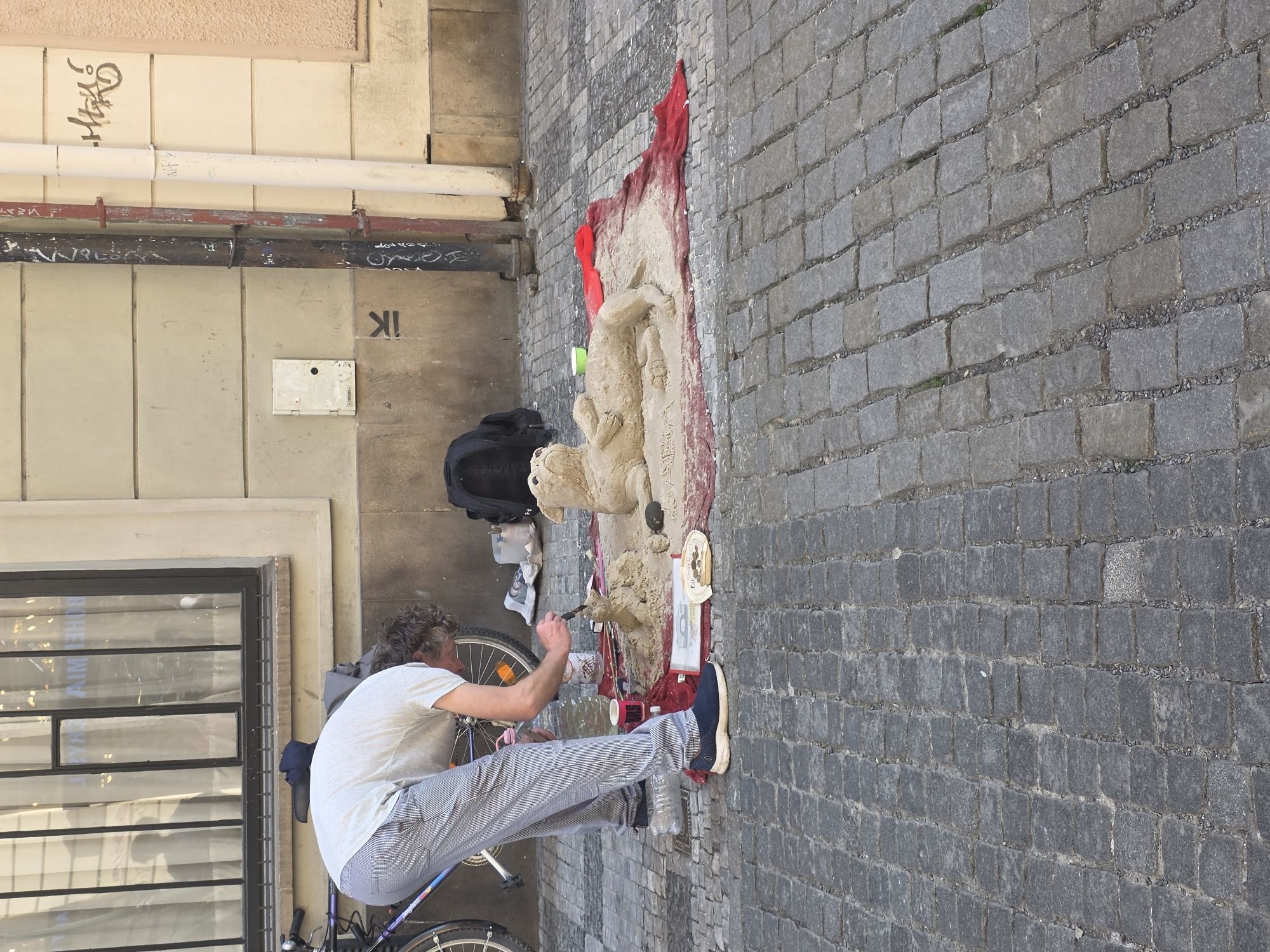 The photograph captures a street artist creating a sand sculpture on a cobblestone sidewalk. The artist, perched on a bicycle seat, is positioned in front of the sculpture, which appears to be an animal figure. He is using a small tool to carve details into the sand. A red cloth is draped behind the sculpture, adding a splash of color to the scene. A backpack and plastic bottles are visible near the sculpture. The backdrop includes a building facade with pipes and graffiti on the walls. The location is Unknown, Unknown.