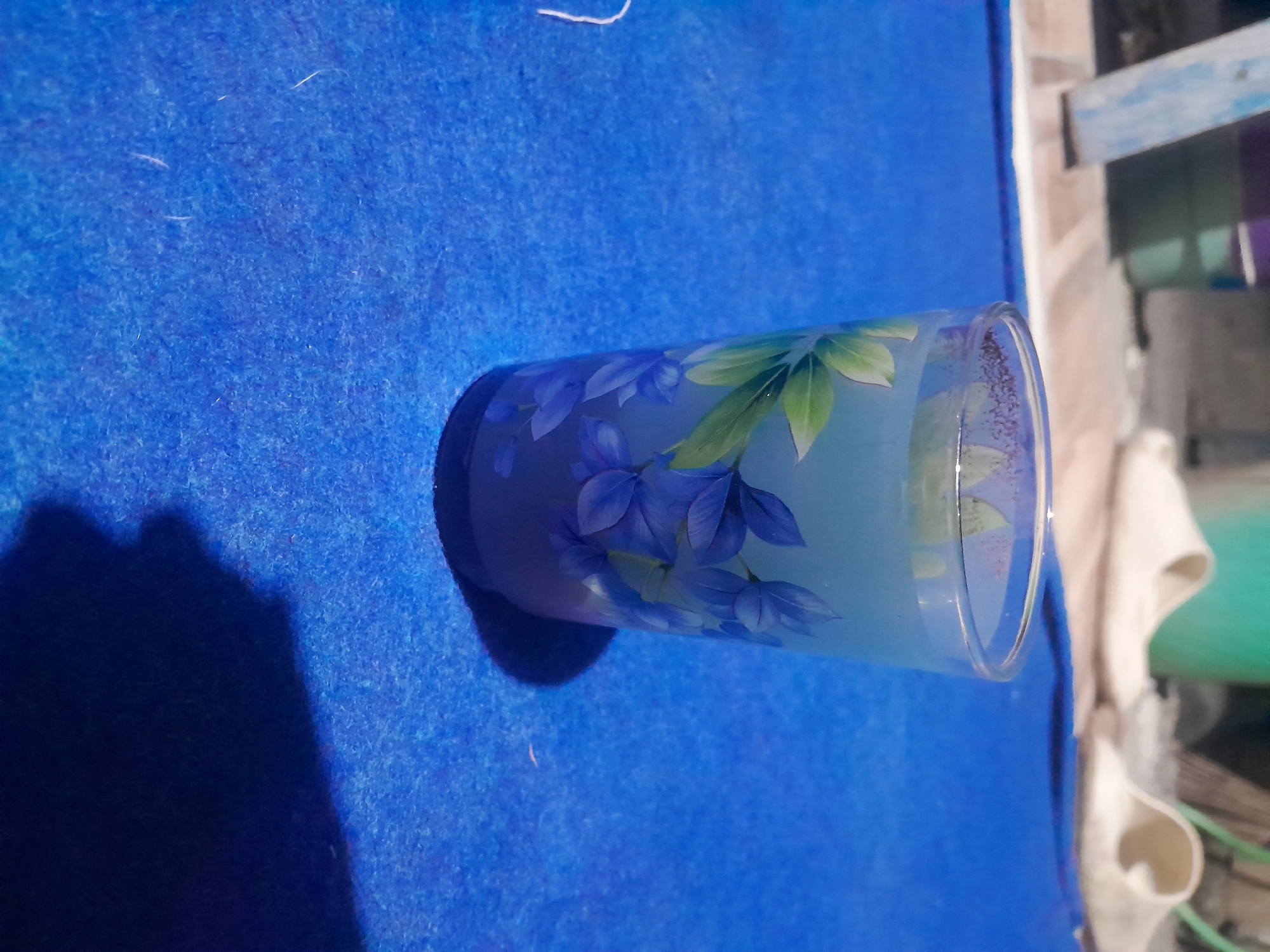 A glass with a floral pattern is placed on a blue surface. The glass contains a light-colored liquid. In the background, there are white slippers and objects that appear to be buckets. The scene suggests an indoor setting, possibly a home, in Pacul, Indonesia, though the specific location within the city is not discernible.