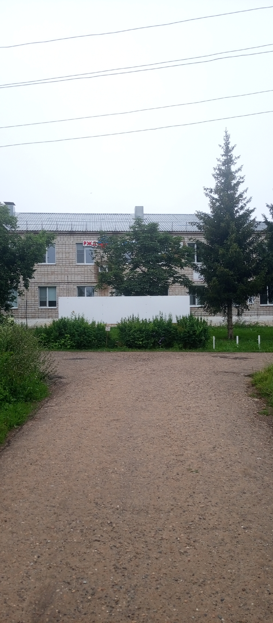 The image captures a cloudy day in Agryz, Russia, featuring a two-story building with a light brown brick facade and a corrugated metal roof. Several windows are visible on the upper floor, some with white frames. A red and white sign with Cyrillic lettering, partially obscured by trees, is mounted above one of the windows. Two chimneys protrude from the roof. In front of the building, a white wall separates it from a row of dense green bushes.
To the left and right of the building are mature evergreen trees, with the one on the right being taller and more prominent. A gravel path leads towards the building from the foreground. The path is bordered by green grass and more dense foliage on the left side. The overall scene appears to be an outdoor area, possibly a public space or the grounds of an institution. There are no people or vehicles visible in the image.