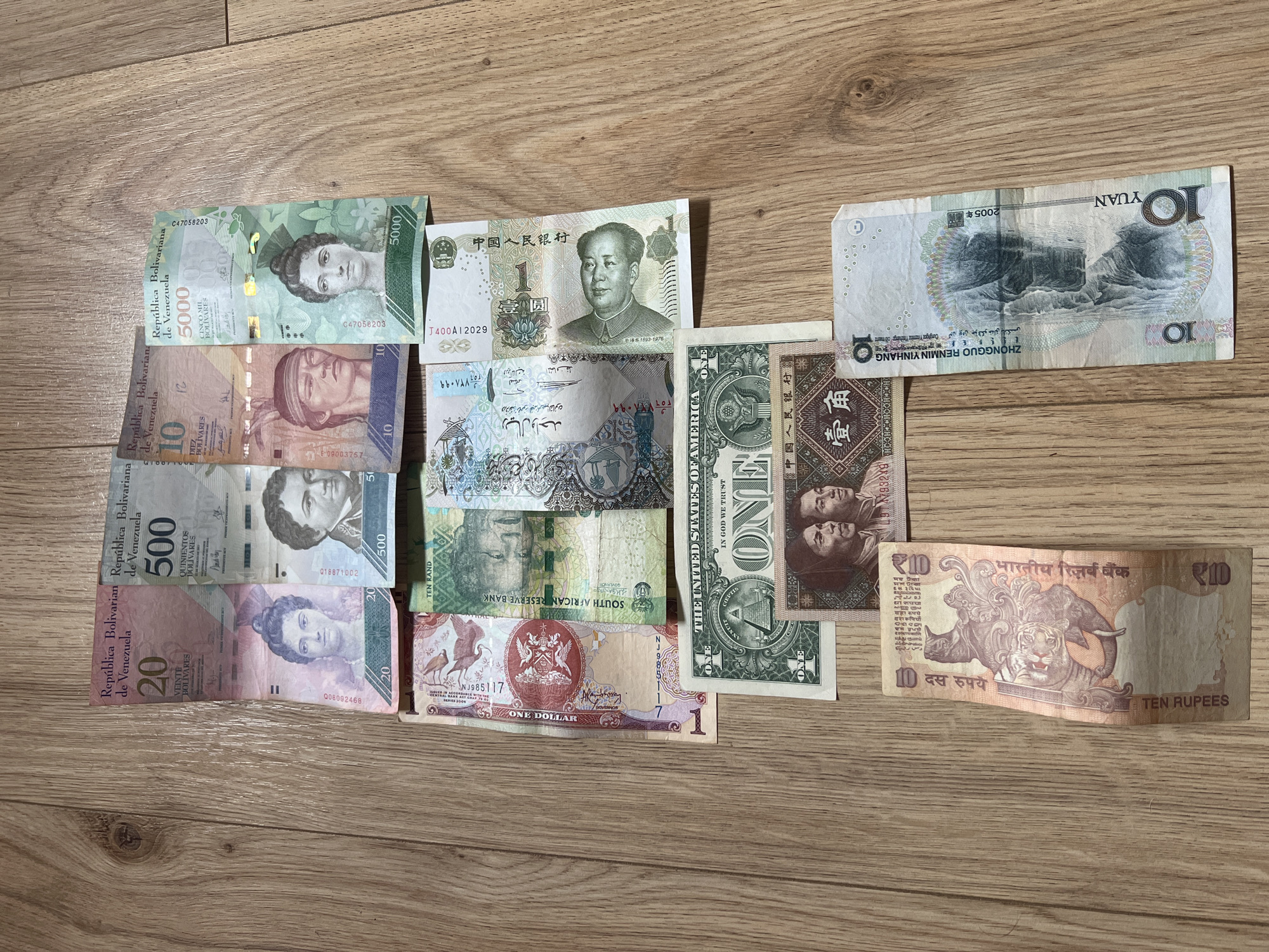 The image shows a collection of various foreign currency banknotes arranged on a wooden surface. There are Venezuelan Bolivares of denominations 20, 500, 10 and 5000; a Chinese Yuan and Jiao; a 10 South African Rand note; a 1 United States dollar bill; a 1 Omani Rial note; a Zimbabwe dollar note; and an Indian 10 Rupee banknote. The bills are overlapping and positioned at varying angles. The wooden surface has a light brown color with visible grain patterns.