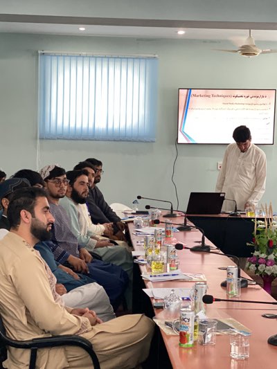 A group of men are seated at a conference table in a room with a presentation being given. The men, dressed in traditional clothing, are looking towards a presenter who stands at the end of the table next to a laptop. A screen displays the title "Marketing Techniques". Microphones and water glasses are placed on the table. The room has vertical blinds on the windows, a ceiling fan, and light-colored walls. A floral arrangement sits on the table near the presenter. The location is Shurandam, Afghanistan.