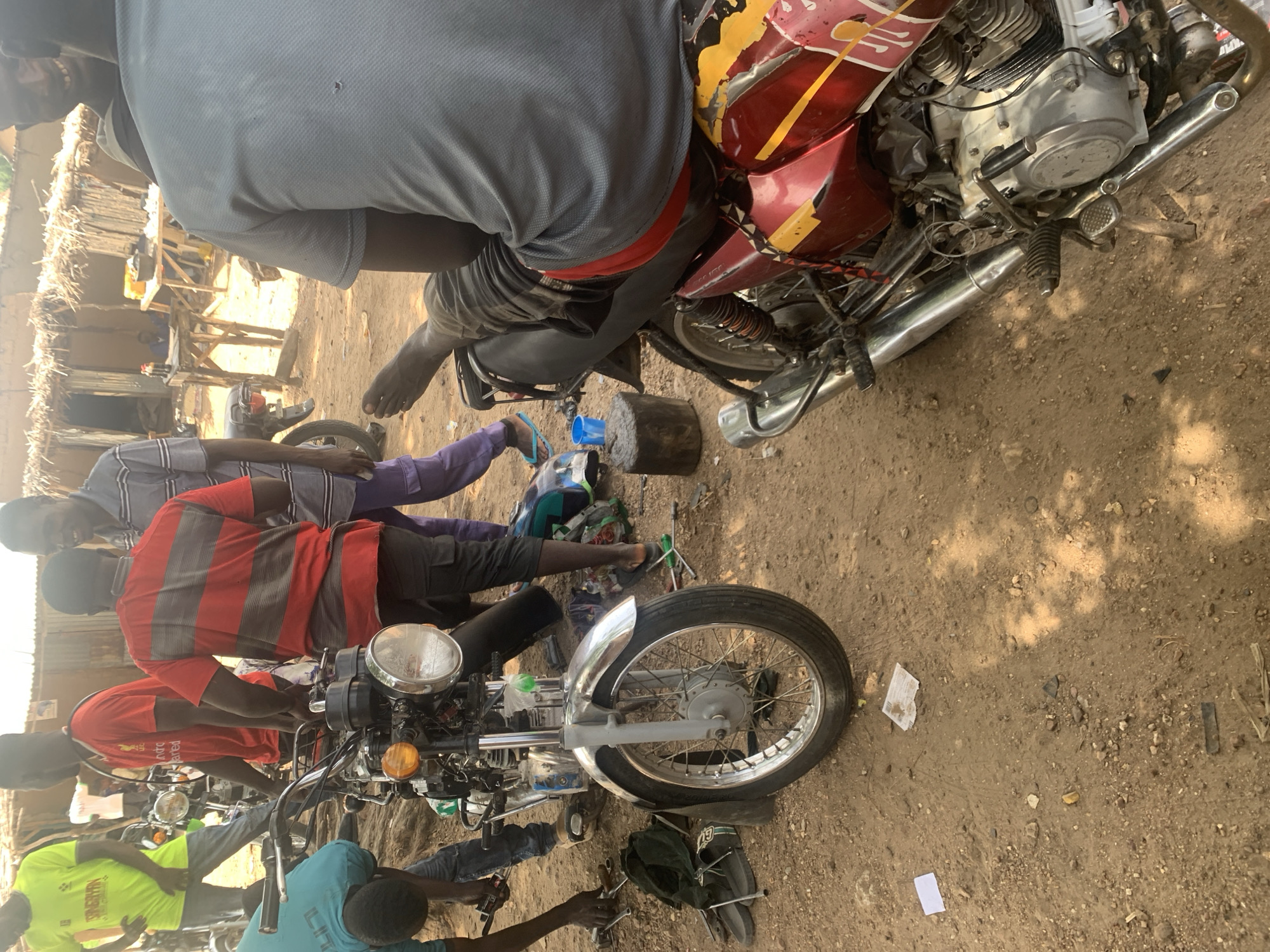 In Gobbiya, Nigeria, a group of men are gathered around two motorcycles in a dusty, outdoor setting. One motorcycle, predominantly silver and red with yellow accents, is in the foreground, viewed from the right side. A person is seated on this motorcycle, with their bare foot resting on the ground and their upper body turned away from the camera. Tools and debris are scattered on the dirt ground around the vehicles.
Behind this motorcycle, another motorcycle, primarily silver with a red seat, is partially visible. Several men are actively engaged with the motorcycles. One man in a bright yellow t-shirt is standing to the left of the silver and red motorcycle, looking down. Another man in a red and black striped t-shirt is standing next to the silver and red motorcycle, appearing to be working on it or observing. A man in a gray and purple striped shirt is standing to the right of the red and black striped t-shirt wearer, looking towards the front. In the background, a simple thatched-roof structure and some wooden tables are visible, suggesting a roadside workshop or market stall. The environment is characterized by dry, sandy earth and sparse vegetation. The overall impression is one of activity and repair work being carried out in a rural or semi-urban Nigerian setting.