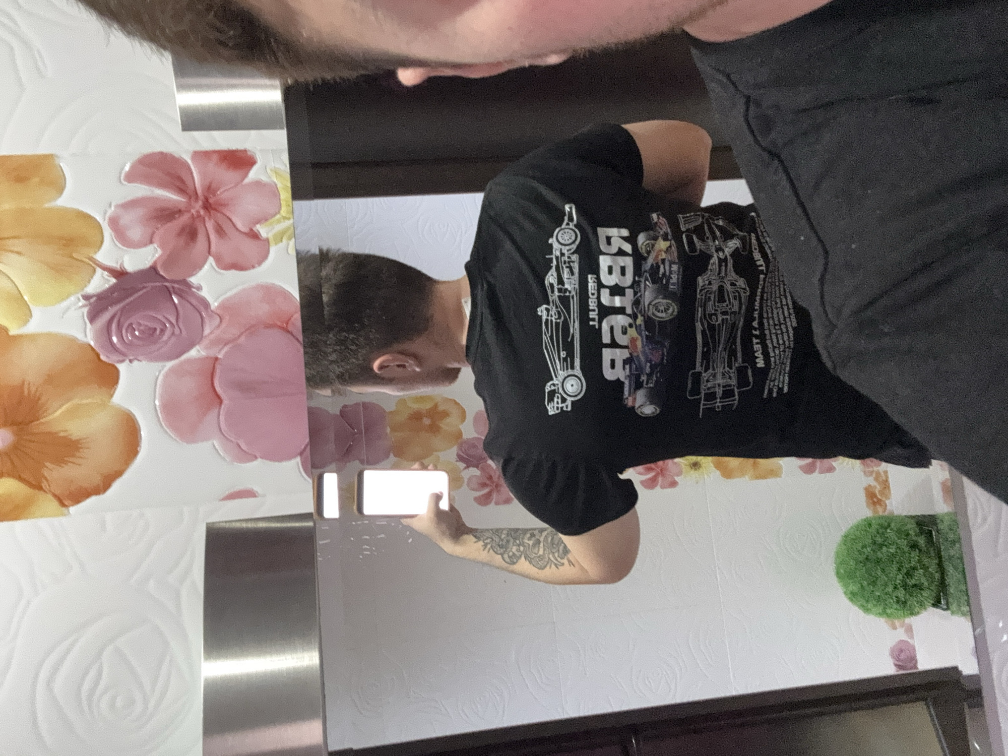 A man is taking a picture of himself in a mirror. He is wearing a black t-shirt with a racing car graphic on it. He is holding a phone in his right hand. His left arm is visible and has a tattoo on it. The background consists of a tiled wall with floral decorations and a small, round green plant. The location cannot be determined from the image.
