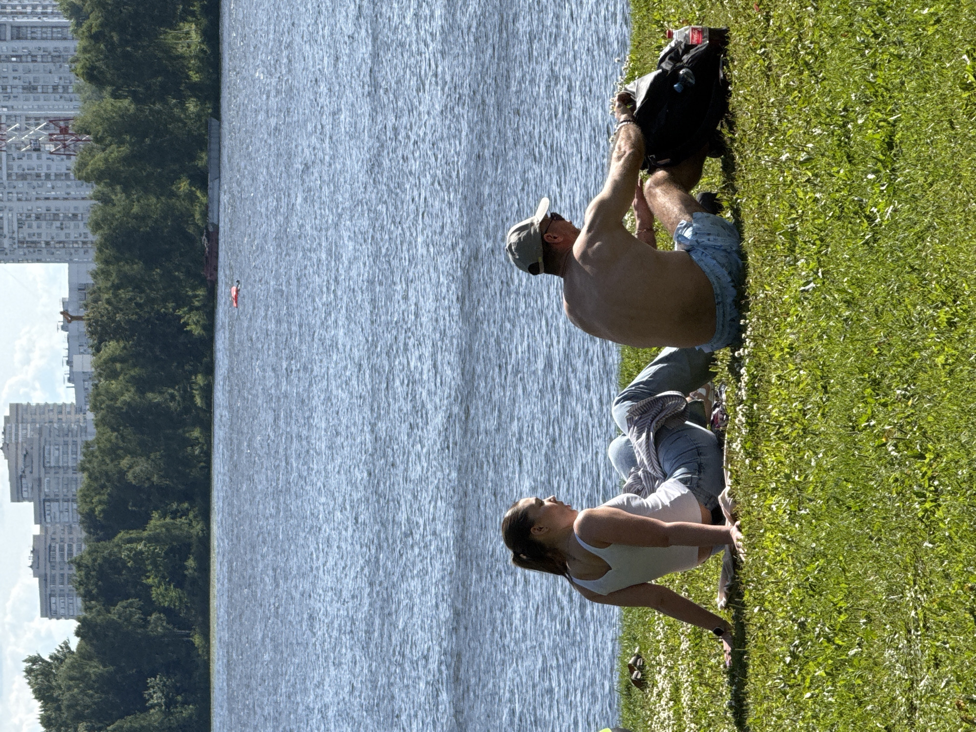 In Khimki, Russia, a man and a woman are sitting on a grassy embankment overlooking a wide body of water, likely a river or reservoir, under a bright, cloudy sky. The woman, positioned on the left, is wearing a white tank top and blue jeans, her legs extended and bent at the knees. She has dark hair pulled back in a ponytail and is looking to her right. A light-colored blanket or towel is spread beneath her on the grass.
To her right, the man, shirtless and wearing blue patterned shorts and a baseball cap, is also seated on the grass. He is leaning forward, with his right arm reaching towards a black backpack placed next to him. A bottle of Coca-Cola rests beside the backpack. The background features a dense line of green trees, beyond which are tall apartment buildings and a construction crane visible in the distance. The water is calm with ripples, and a small red object, possibly a life raft or buoy, is visible on its surface. The scene suggests a relaxed outdoor recreational setting.