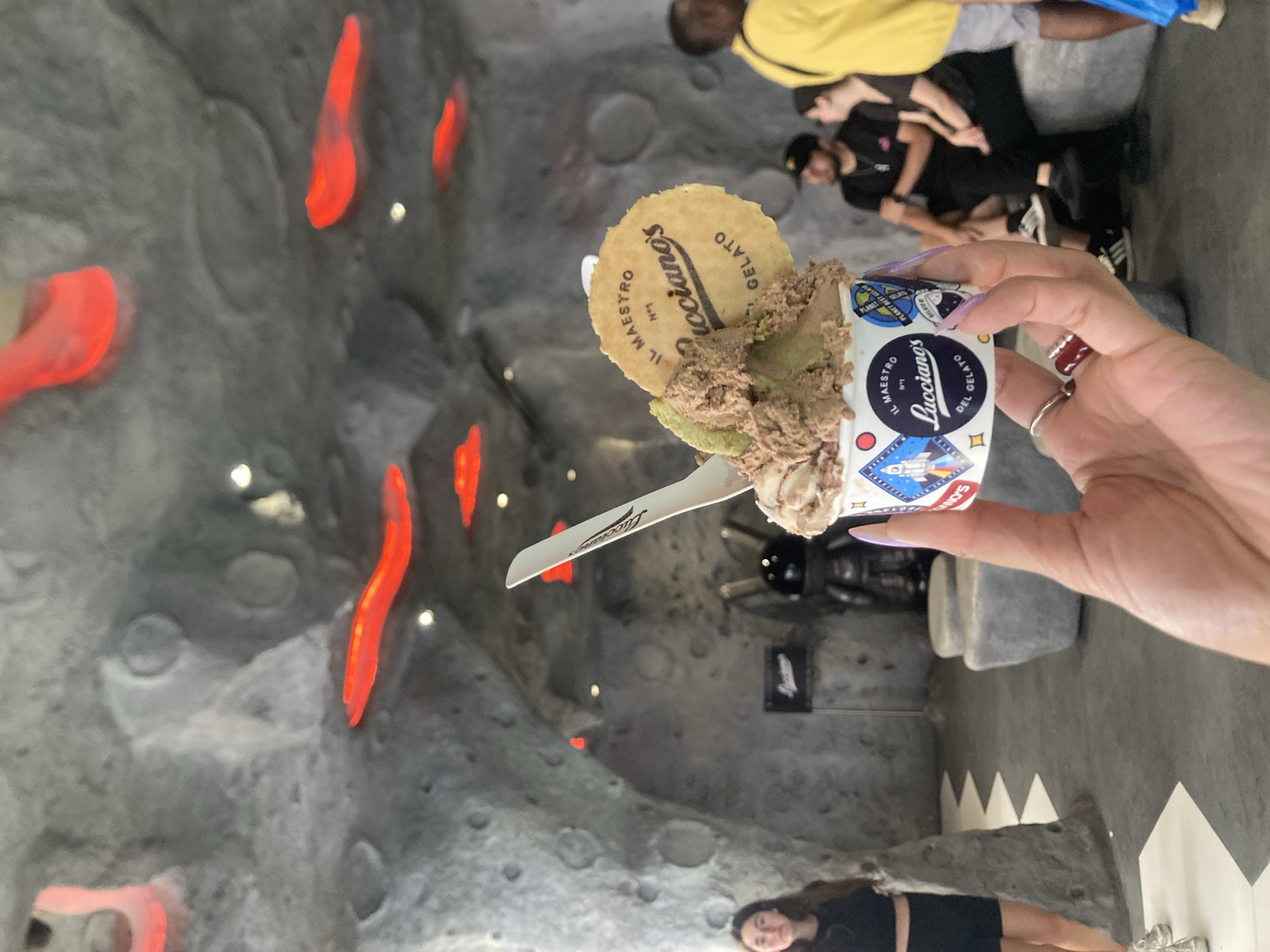 This media file presents an indoor scene within a gelato shop, characterized by its distinctive, dark, and textured walls resembling a cave or lunar landscape. Several bright red, organic-shaped light fixtures are embedded in the walls, alongside smaller white spotlights, contributing to a unique ambiance.
In the foreground, a hand with long, light purple fingernails and a red-jeweled ring holds a cup of gelato. The gelato itself appears to be a swirl of brown and green flavors, possibly chocolate and pistachio, and is topped with a round, decorative waffle. The waffle bears the text "IL MAESTRO N°1 Lucciano's GELATO." The white plastic spoon emerging from the gelato also features the brand name "Lucciano's." The side of the gelato cup displays a graphic of a rocket ship and stars, along with the text "IL MAESTRO N°1 Lucciano's DEL GELATO" and "PLANET BEST GELATO." The word "SONG" is partially visible near the top of the cup.
Several other individuals are visible in the background, out of focus, dressed in casual attire. One person with long dark hair is seen looking towards the camera in the lower right corner, wearing a black top and skirt. Others are standing further back, and some seating or structural elements that resemble large, rough-hewn stones are also present. The scene depicts a casual visit to a gelato establishment, specifically Lucciano's, which is located in Barcelona, Spain. The time of day is not evident, but the setting is clearly indoors with artificial lighting.