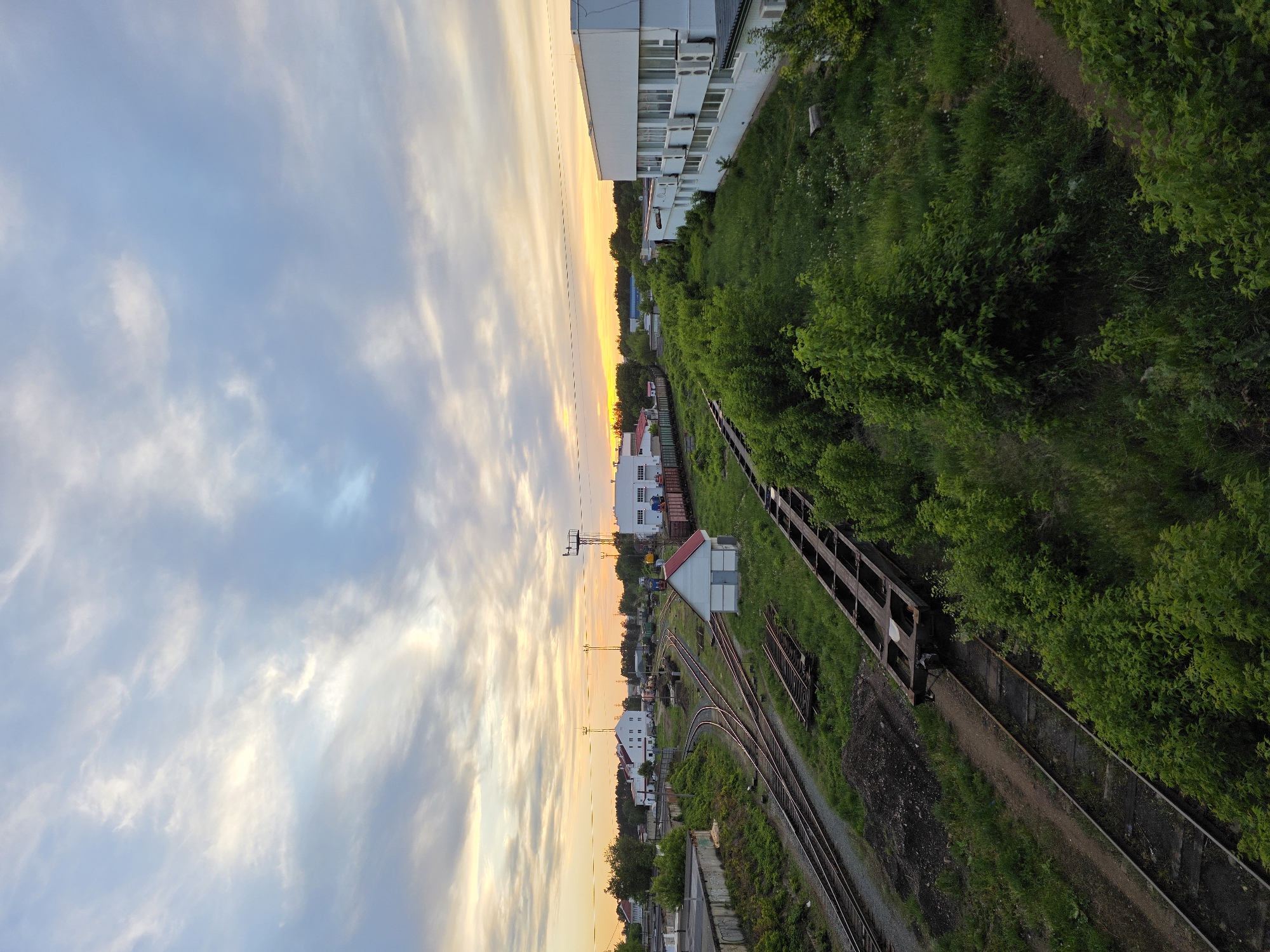 An elevated, angled aerial photograph depicts a section of the city of Chaikovsky, Russia, at twilight. Dominating the scene are railway tracks running alongside dense green vegetation and a concrete structure. A building with visible air conditioning units sits atop a green area. Several small structures and houses are visible further down the railway tracks. The sky is a mix of blue and orange hues, indicating either sunrise or sunset. The image presents a combination of urban elements and natural landscapes, highlighting the city's infrastructure against the backdrop of a colorful sky.