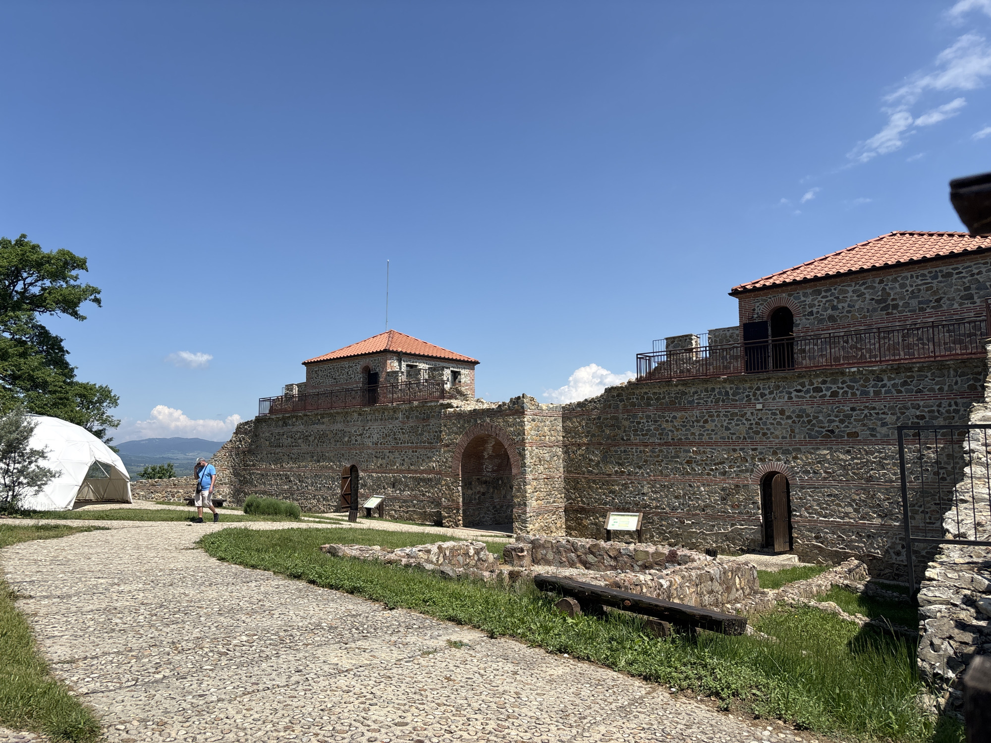 The image captures an outdoor scene at a historical site within Belchinski Bani, Bulgaria, under a clear blue sky with sparse white clouds. The central focus is a series of stone-built structures resembling a fortress or renovated historical complex. These structures are characterized by walls constructed from irregularly shaped grey and brown stones, interspersed with horizontal bands of red brick and mortar.
Two distinct buildings with pitched roofs, covered in terracotta-colored tiles, are prominent. The building on the left features a single antenna extending upwards from its roof. Both buildings have arched openings and dark metal railings along their upper perimeters, providing balcony or walkway access. A large, arched gateway is visible within the main stone wall, suggesting an entrance. Multiple smaller, dark wooden doors with arched tops are set into the stone walls at various points.
In the foreground and midground, a light-colored, cobblestone path curves from the lower left towards the center and then recedes to the left. Lush green grass borders the path, interspersed with smaller stone foundations and remnants of structures. To the left of the path, a large, white geodesic dome-shaped tent is situated next to a leafy green tree. Further down the path, a male individual walks away from the viewer towards the left. He wears a light blue short-sleeved collared shirt, white shorts, and dark shoes, and carries a dark bag over his right shoulder.
To the right of the path, a dark wooden log or beam rests horizontally on the grass amidst some low stone remnants. Two white informational signs on dark posts are positioned near the architectural features. In the distant background, rolling green mountains are visible beneath the bright, sunny sky. The overall scene is well-lit by natural daylight.