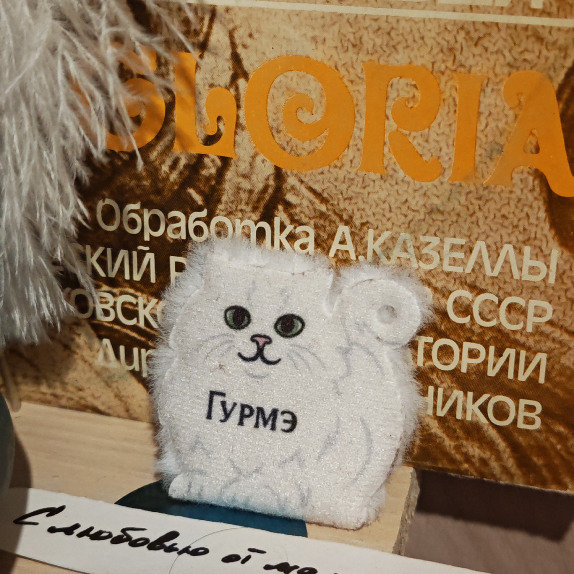 The image captures a close-up still life featuring a white, fluffy, round plush toy resembling a cat, set against a blurred background of an old book or album cover.
The central subject is the white plush toy, which has simple features: small, dark green eyes, a pink triangular nose, and a curved black line for a mouth, giving it a friendly expression. Two small, round ears are visible at the top. The word "Гурмэ" (Gourmet) is printed in black uppercase letters on its front. The toy rests on a light wooden surface, with a dark blue element partially visible beneath it.
In the background, a brown-toned object, possibly a record sleeve or book cover, features large orange text at the top, which appears to spell "GLORIA", though partly obscured by soft, white feathery material on the left. Below this, various lines of white Cyrillic text are visible. These include "Обработка А. КАЗЕЛЛЫ" (Arrangement by A. KAZELLY), "CCCP" (USSR), and partial words like "КИЙ Р", "ОВСК", "ТОРИИ", and "НИКОВ", suggesting titles or credits related to Soviet-era musical or literary works.
Below the plush toy, a white piece of paper with handwritten Cyrillic text is partially visible, starting with "С любовью от" (With love from), with the rest of the message cut off by the bottom of the frame. The scene appears to be an indoor arrangement. The presence of Cyrillic text, particularly "CCCP" and the location context of Kladovitsy, Russia, suggests the items are from or related to Russia.