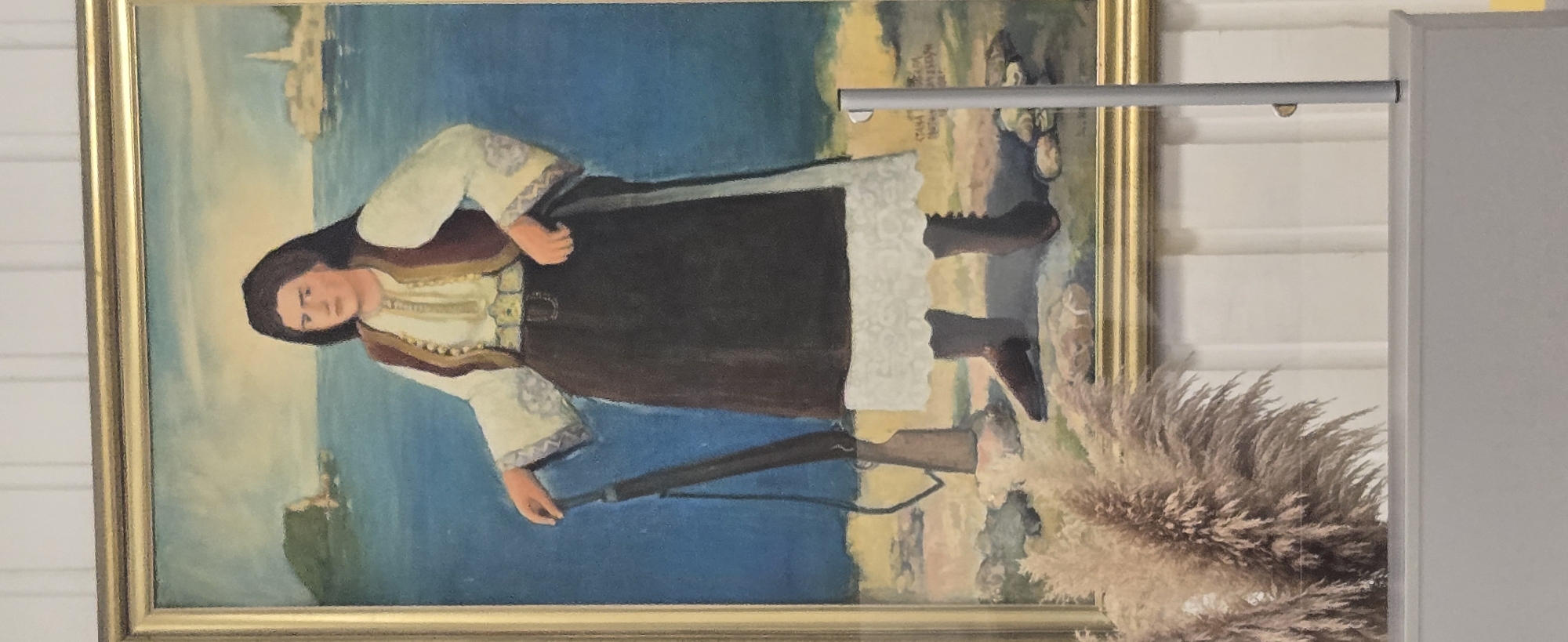 The image shows a framed painting depicting a woman holding a rifle. She wears traditional attire with a white and brown color scheme. The painting's background portrays a coastal scene with blue water and a land formation that includes a building or structure suggestive of architecture. The frame of the painting is gold. The scene in the painting resembles the landscape near Budva, Montenegro, with its characteristic coastline and historical buildings, potentially depicting a Montenegrin woman holding a firearm as part of local resistance history. The painting is partially obstructed by modern elements like a grey support structure and a decorative feather plume.