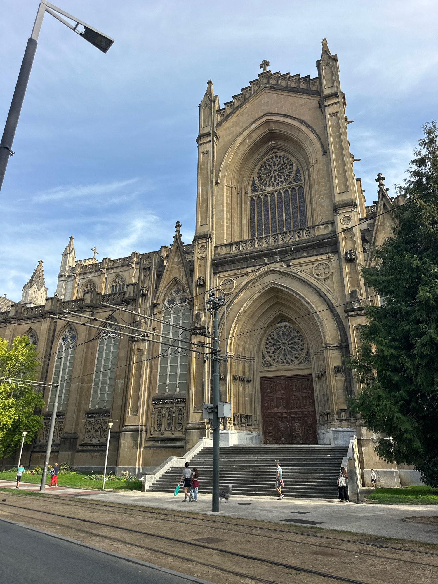 The image shows the facade of the Cathedral of Mary Immaculate in Vitoria-Gasteiz, Spain. The cathedral is a large Gothic Revival building constructed of light-colored stone. The main entrance is a large, arched doorway with double wooden doors. Above the entrance is a rose window, and above that, a large stained-glass window. The facade is flanked by tall spires and decorated with intricate carvings.
In front of the cathedral, a wide set of stone steps leads up to the entrance. Several people are visible on the steps and in the plaza below. On the left side of the image, a streetcar track runs along the sidewalk, with a streetcar visible in the distance. A lamppost stands on the left, and a tall evergreen tree is on the right side of the image. The sky is blue with scattered clouds.