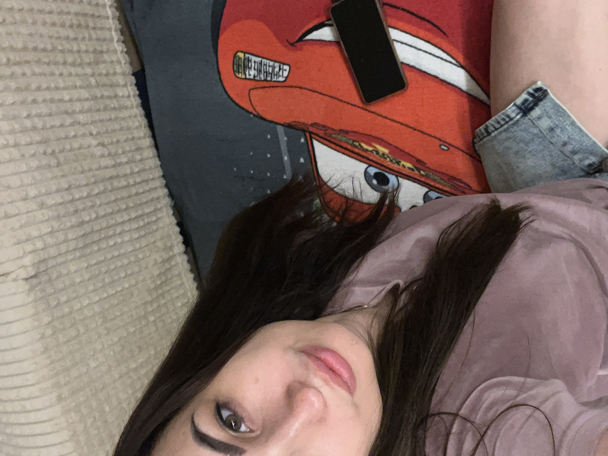 An overhead, eye-level shot shows a young woman from the chest up. She has long, dark brown hair that falls over her shoulder and chest. She is wearing a light pink t-shirt and denim shorts. Her skin is fair, and she has makeup on her face. She is smiling slightly, and her eyes are looking at the camera. She is lying down on a couch with a blanket. The blanket is red and has a picture of Lightning McQueen from the movie Cars on it. A cell phone is lying on top of the blanket next to her hand. The lighting in the room is dim, and there is a slight glare on her face. The background is a blurred out couch.