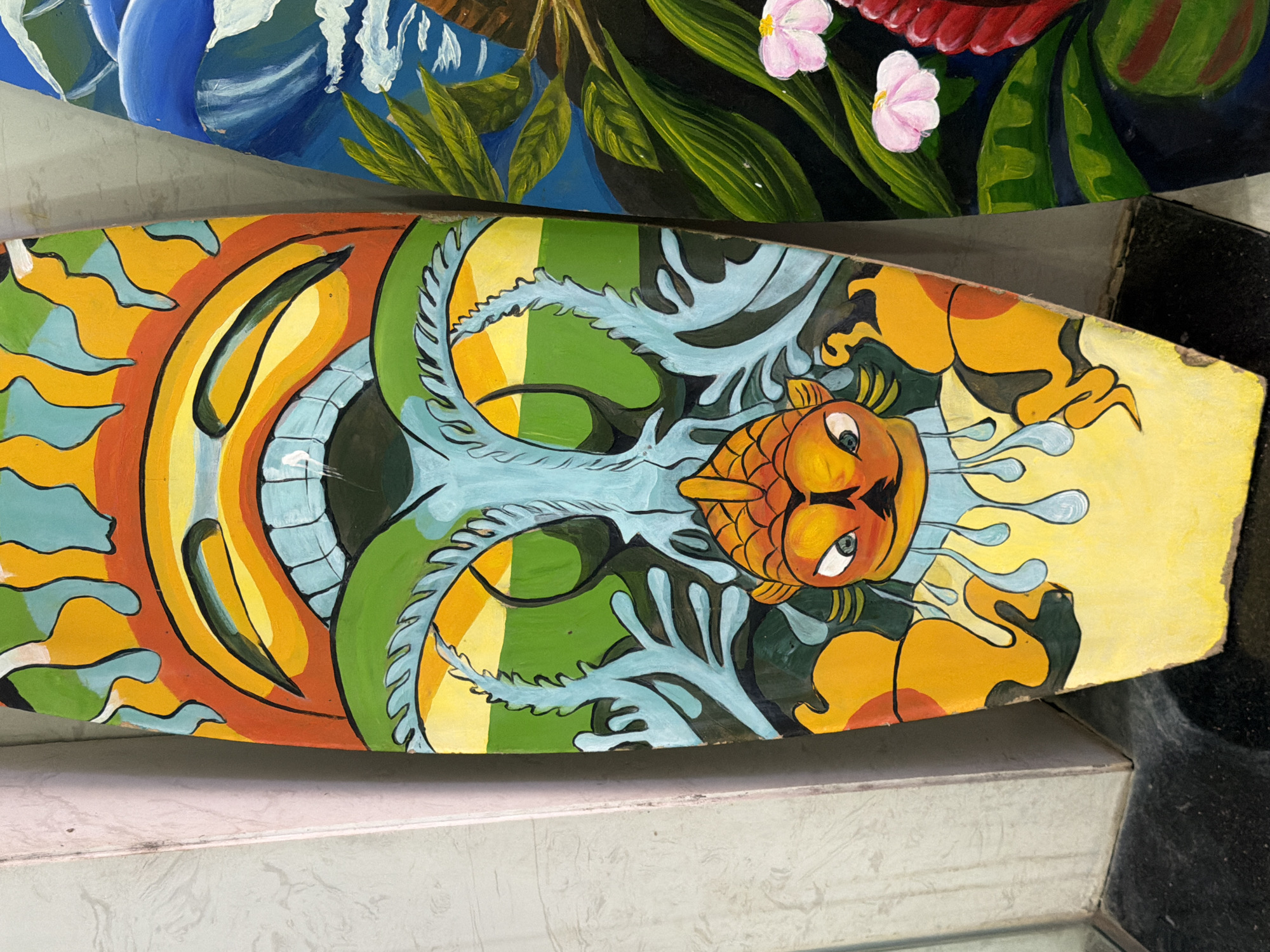 The image displays two decorative surfboards leaning against a wall. The surfboard in the foreground is painted with a vibrant, colorful design featuring a stylized sun with a face at the top, followed by a tree-like structure with flowing branches and a central fish head with large, expressive eyes. The lower portion of this surfboard is a bright yellow. The surfboard to the right is partially visible and features a tropical scene with large green leaves, pink flowers, and a red flower. The surfboards are positioned indoors, near a wall that appears to be painted a light color and a dark floor. The lighting suggests an indoor environment, possibly a shop or gallery. The image does not provide any specific information about its location within the city of Đà Nẵng, Vietnam.