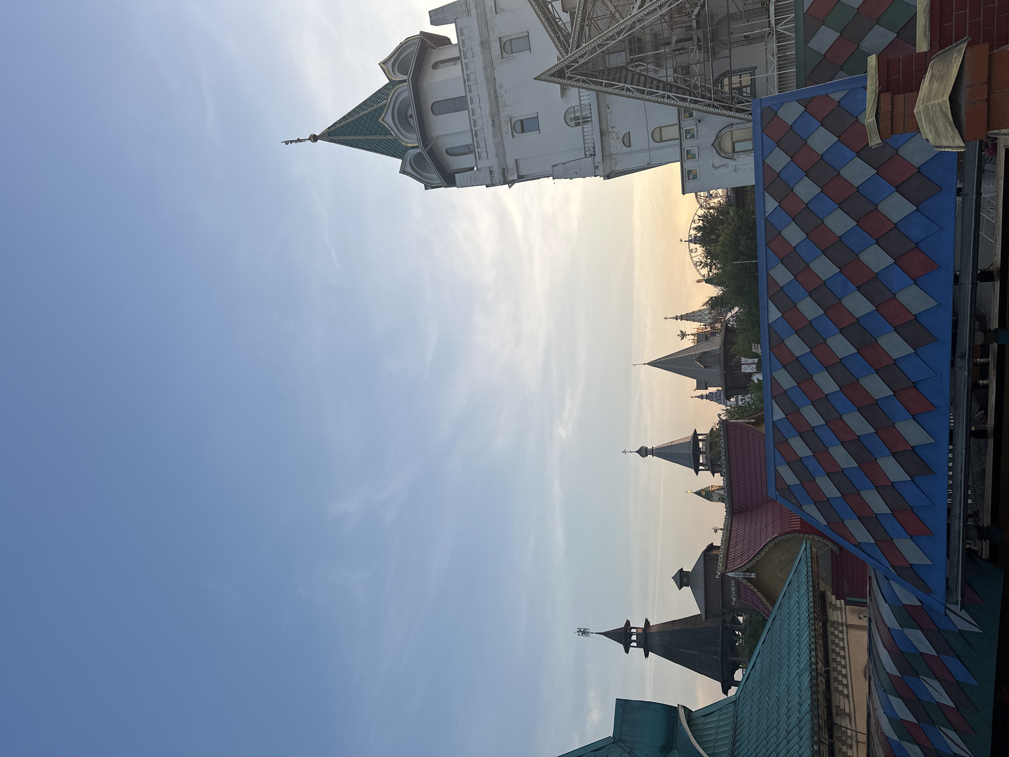 The image presents an elevated, late afternoon or early evening view of the Izmaylovo Kremlin in Moscow, Russia. The sky, transitioning from light blue overhead to a warm, soft orange-yellow near the horizon on the right, exhibits scattered wispy clouds and faint contrails.
On the far right, a prominent white building features a tall, multi-tiered tower capped with a green, conical roof adorned with ornate gold-colored elements and dormer windows. Below this tower, a triangular metal scaffolding-like structure is visible.
The mid-ground reveals a collection of buildings with traditional Russian architectural elements, including numerous dark-roofed turrets and spires, many topped with finials resembling crosses or other decorative motifs. These structures feature steeply pitched roofs with intricate patterns, including one in the foreground with a diamond checkerboard design in blue, red, and gray. Another brightly colored turquoise-green tiled roof is visible on the left. A reddish-brown roof with scalloped edges is seen towards the center-left.
In the immediate bottom-right corner, a brick chimney with a brass-colored cap is partially visible. Further in the distance, behind a cluster of trees and buildings, the upper segment of a Ferris wheel or similar amusement ride is faintly discernible against the horizon's glow. No individuals are present in the scene.