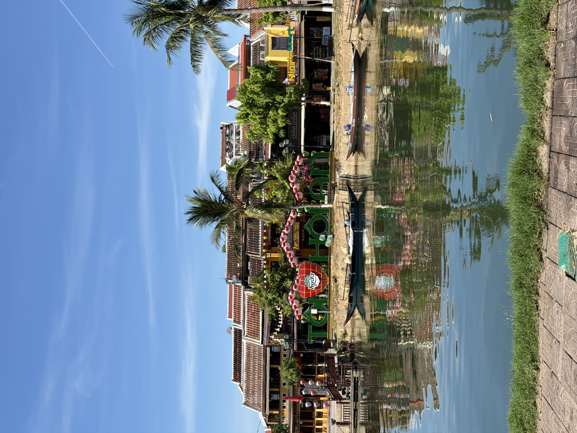 This is a photograph of the Thu Bon River in Hội An Ward, Vietnam, taken on a sunny day. The river reflects the colorful buildings and lush greenery lining its banks. Traditional Vietnamese architecture, characterized by yellow facades and tiled roofs, is visible along the riverfront. A large, illuminated heart-shaped sign with the words "TO HOI AN" and "HUETU HUETU" is prominently displayed. Several boats, some carrying people, are moored along the river, while others appear to be in motion. Palm trees and other vegetation add to the tropical ambiance of the scene. The sky is clear blue with a few wispy clouds, and a faint contrail from an airplane is visible. The foreground features a grassy embankment and a paved walkway.