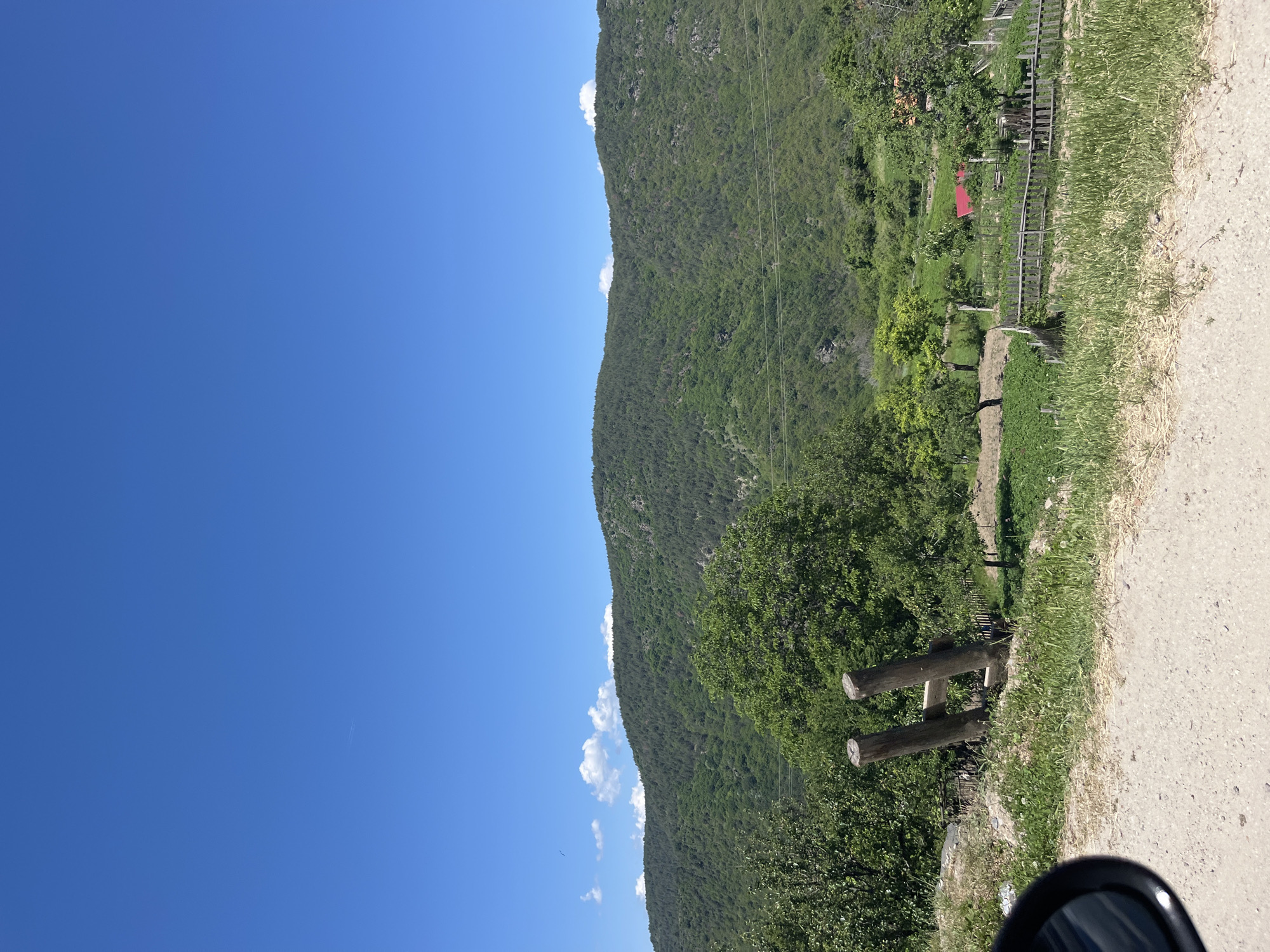 The image shows a landscape in Leshten, Bulgaria, with a clear blue sky and a tree-covered mountain. A road runs alongside the mountain. A wooden structure with two horizontal logs stands next to the road. Some houses with red roofs are visible on the mountainside. The edge of a car mirror appears in the bottom right corner.