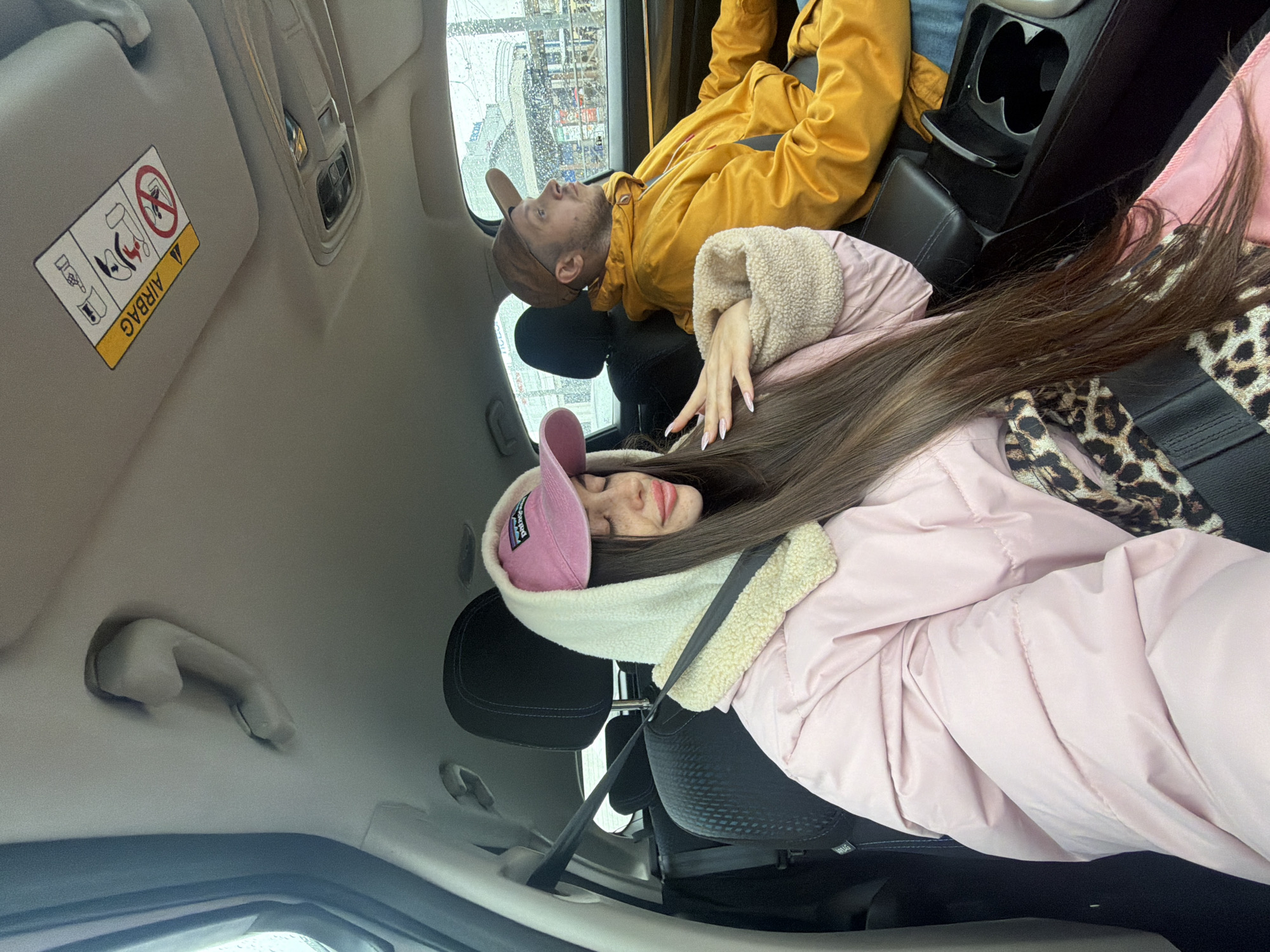 The image captures a couple inside a car, likely in Saint Petersburg, Russia, given the urban landscape visible through the car window. The woman, wearing a pink cap and a light pink jacket with a cream-colored fluffy hood, is reclined in the passenger seat. Her long brown hair is spread out. She is wearing a seat belt. Next to her, the man is in the back seat, wearing a brown cap and a yellow jacket. He is also reclined. The car's interior is visible. Details such as the airbag warning label, a grab handle, and the car's controls are present. Outside, there is a building. The weather appears overcast or rainy.