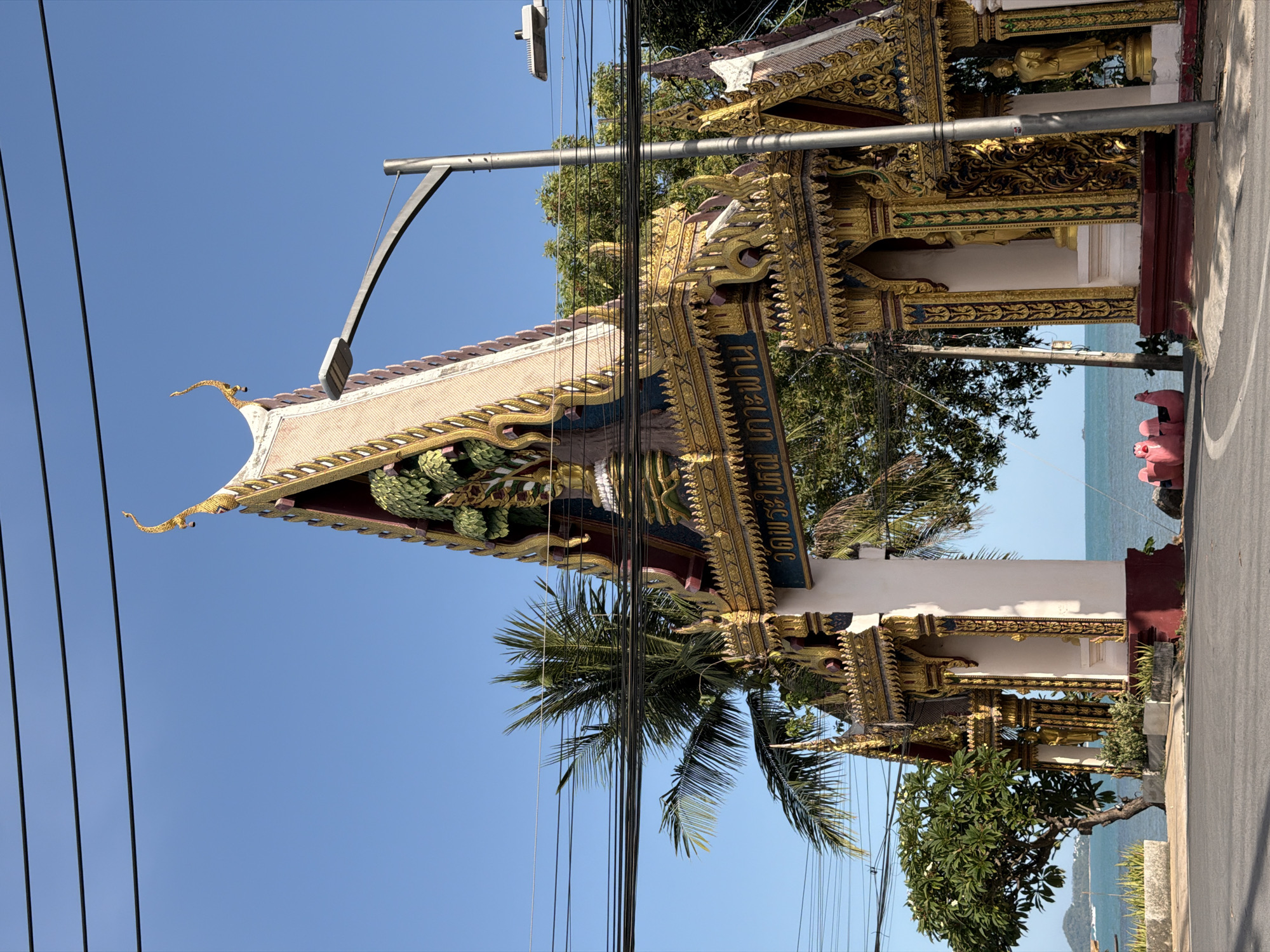 The image depicts a grand, ornate archway in the city of Ko Samui District, Thailand. The archway is decorated with intricate golden carvings and features Buddhist imagery. Behind the archway, lush green trees and a palm tree are visible, with the turquoise ocean and a clear blue sky forming the backdrop. The scene is bordered by power lines stretching across the top of the frame. There are no people visible in the image.